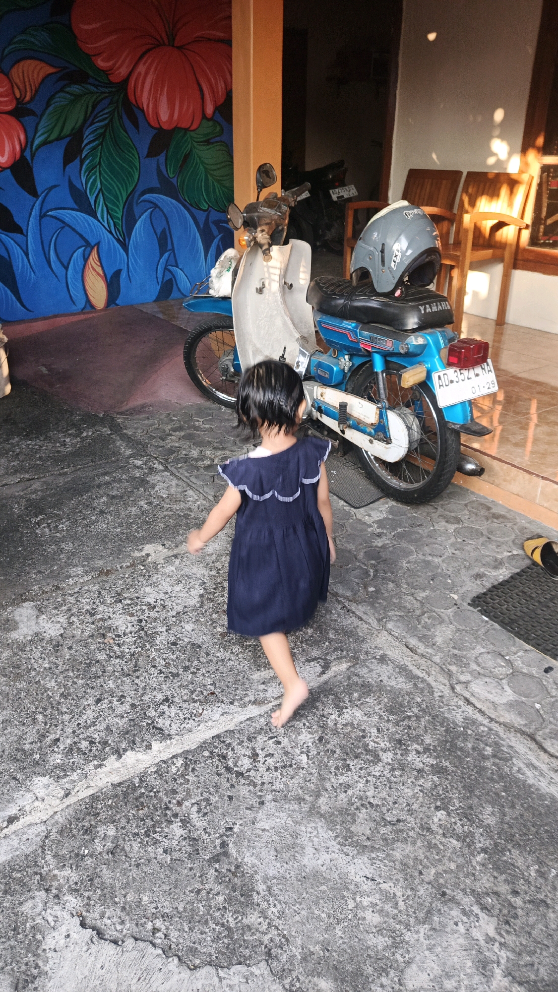 The image depicts a young child, viewed from behind, walking barefoot on a textured concrete pathway. The child is wearing a dark blue sleeveless dress with a white ruffled trim. Adjacent to the child is a light-colored motorcycle with a blue fender and a black seat. A grey helmet rests on the motorcycle's seat. In the background, a mural with large red flowers and blue foliage adorns a wall, alongside an orange pillar and two wooden chairs. The setting appears to be an outdoor area, possibly a courtyard or driveway, with the architecture suggesting a location in Surakarta, Indonesia. The lighting indicates it is daytime, with sunlight casting shadows.