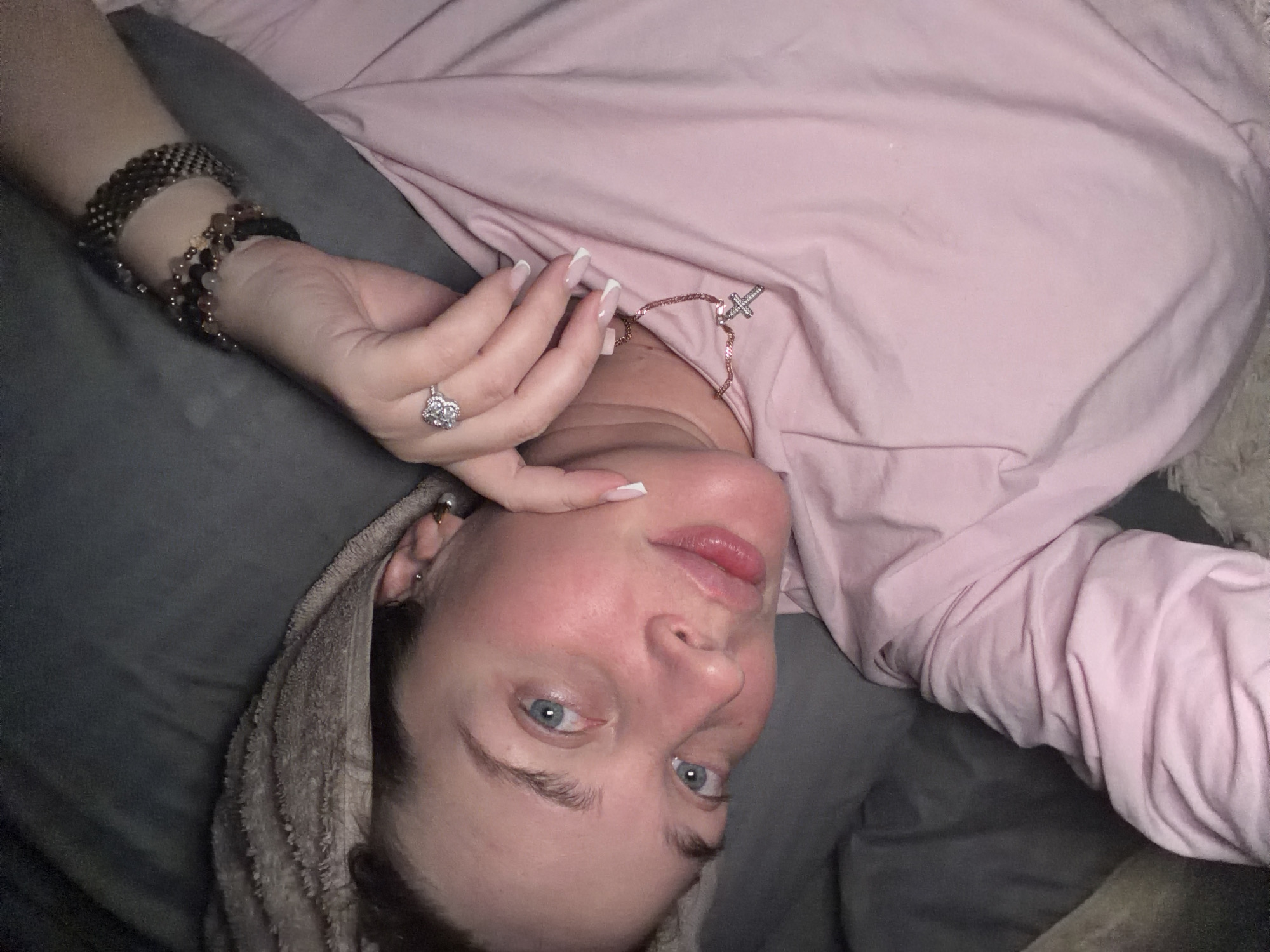 A person is lying down, facing the camera. The individual has fair skin, blue eyes, and is wearing a pink long-sleeved shirt. A gray towel is wrapped around their head. On their left wrist, they are wearing multiple bracelets, and a large ring is visible on their left hand. A necklace with a cross pendant is around their neck. The person is lying on a gray pillow or cushion. The image is cropped and angled. There is no evidence to suggest this image was taken in Chaikovsky, Russia.