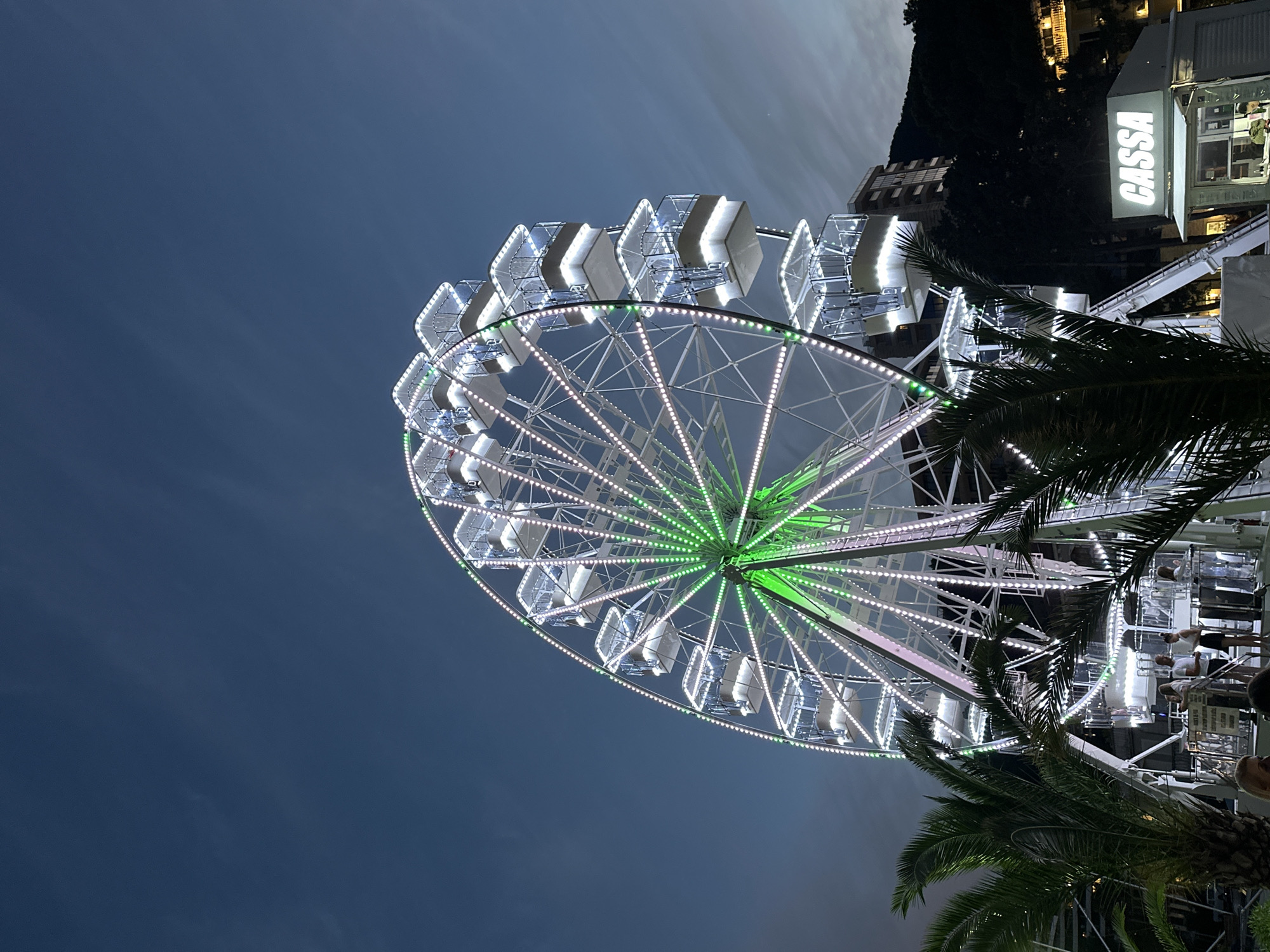 This image depicts a Ferris wheel illuminated with white and green lights at dusk in Budva, Montenegro. The Ferris wheel is centrally located, dominating the frame with its intricate structure. Several cabins, lit from within, are visible as they ascend the wheel. In the foreground, palm trees frame the scene, their fronds partially obscuring the base of the Ferris wheel. To the right, a small booth with the illuminated sign "CASSA" is present, suggesting a ticket or information counter. Behind the Ferris wheel and to the right, a multi-story building and a hillside covered with trees are discernible. The sky is a deep blue, indicative of twilight, with faint cloud formations. A few individuals are standing near the base of the Ferris wheel and by the "CASSA" booth, engaged in casual interaction. The overall atmosphere suggests a recreational setting, possibly an amusement park or a waterfront attraction.