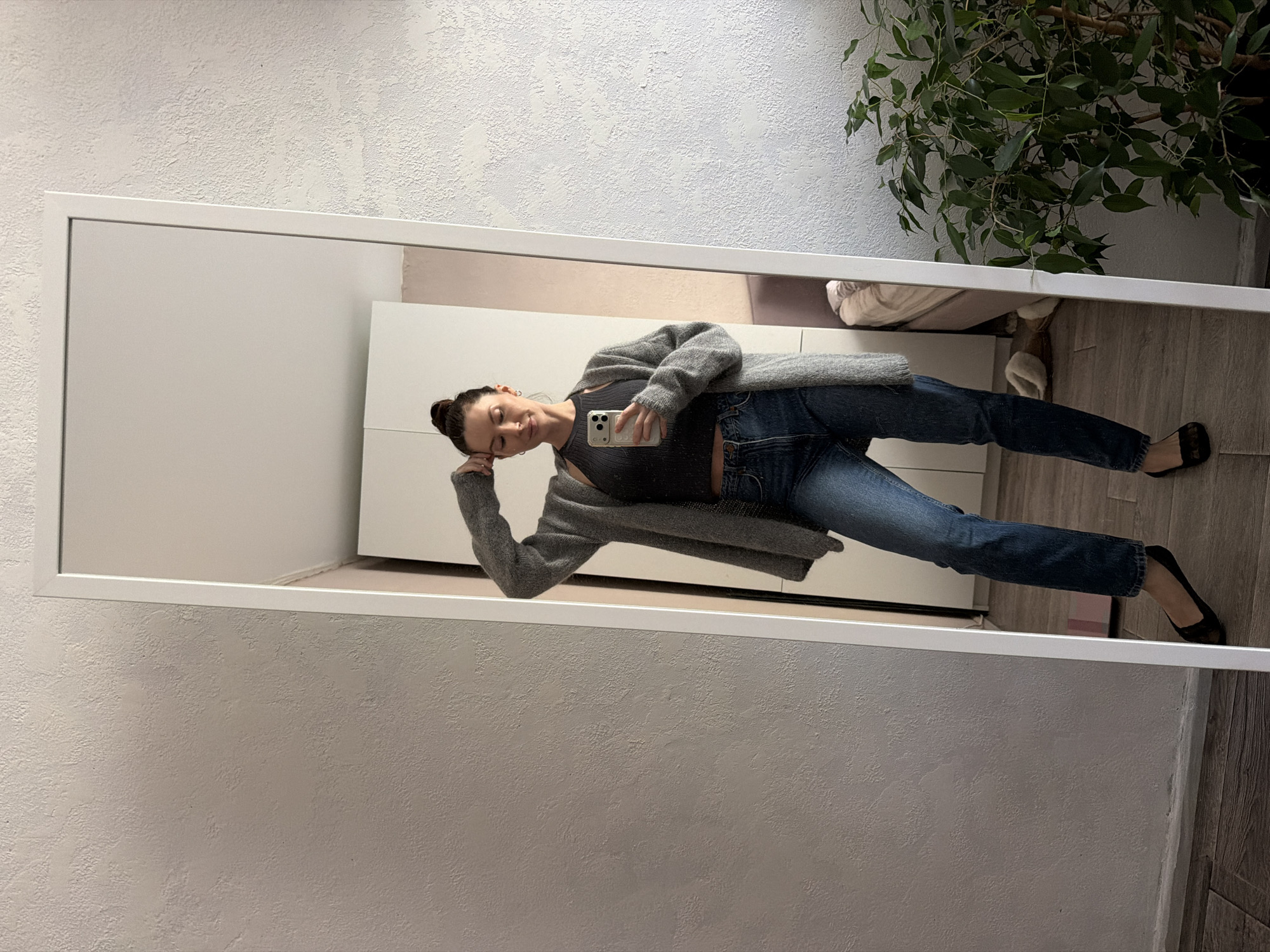 A woman stands in front of a mirror, holding a phone in her hand and leaning her head on her other hand. She is wearing a dark tank top, blue jeans, a gray cardigan, and black flats. The mirror is framed in white and is positioned in a room with light gray walls, a white cabinet, a plant, and a wooden floor. The exact location in Perm, Russia cannot be determined from the image.