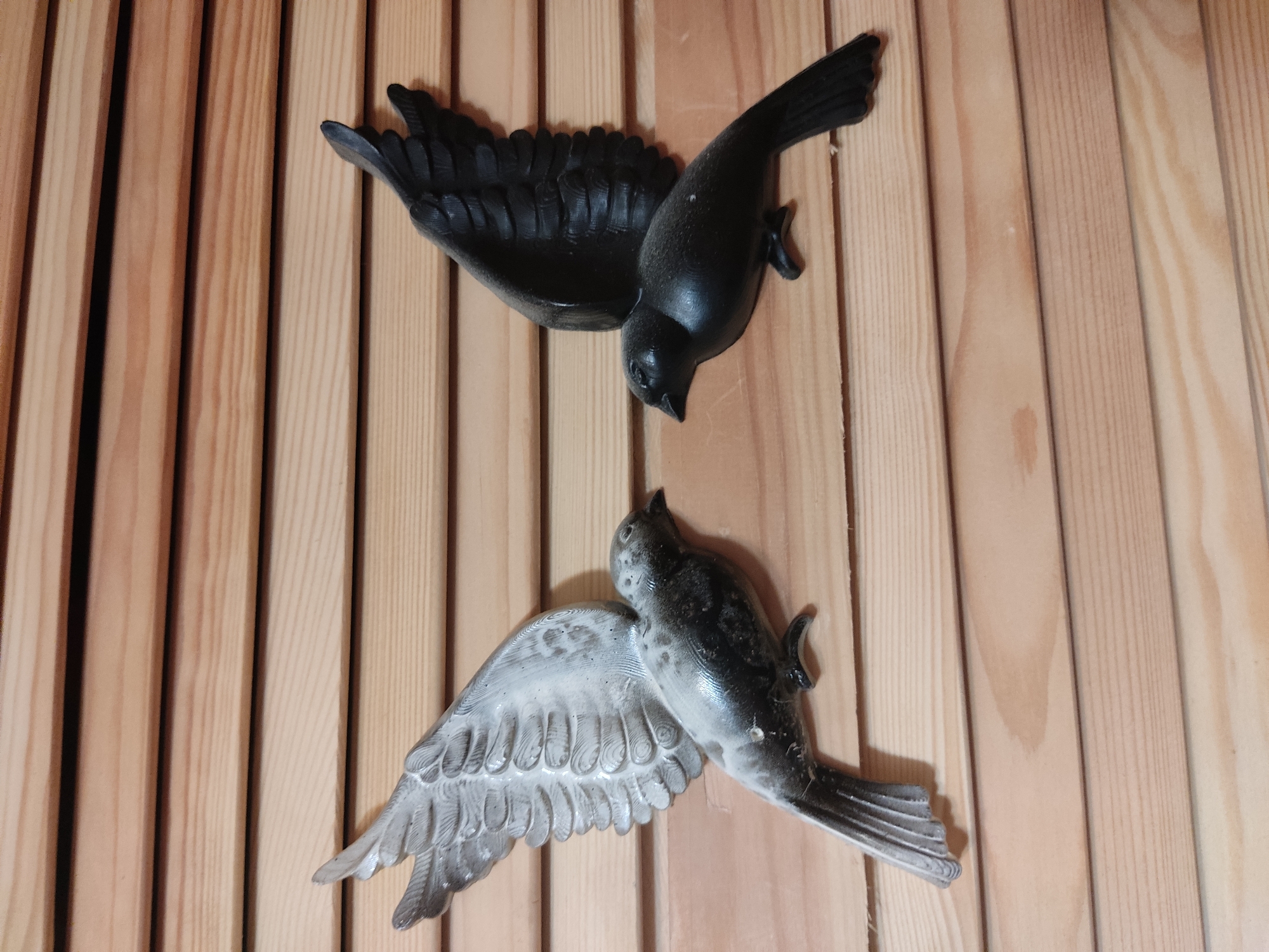 Two decorative wall-mounted bird figures are displayed on a horizontal wooden slat wall. The bird on the left is predominantly silver with dark accents, and its wings are spread upwards. The bird on the right is entirely black, with its wings also spread upwards. The background consists of light-colored wooden planks with visible grain. The scene appears to be indoors, possibly in a home or shop. There is no information in the image to determine its location in Noginsk, Russia.