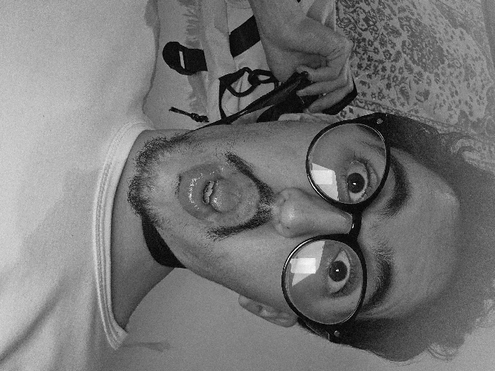 A person, seemingly male, lies on their back, face tilted towards the camera. They wear large, round glasses. The mouth is open, tongue extended. A beard is visible. A hand holds a device above the head. Clothing details include a light-colored t-shirt and a patterned fabric in the background. The image is in black and white.