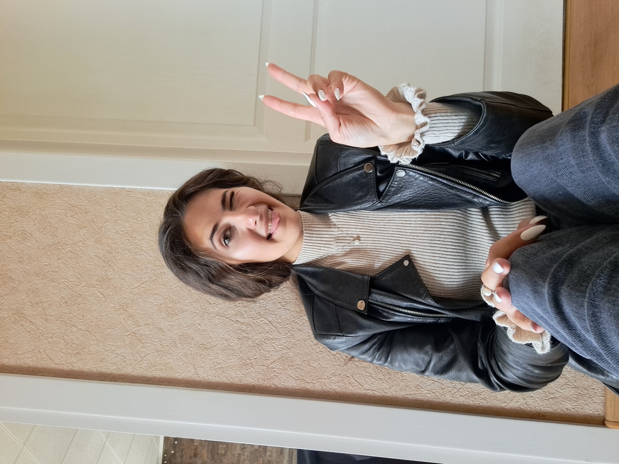 A young woman poses indoors. She is wearing a light-colored, horizontally striped top with a button at the neck, and a black leather jacket. She is also wearing blue jeans. Her left hand is raised making a peace sign. She is winking and sticking her tongue out slightly. The background includes a beige textured wall on the left, a white door in the upper right, and a white frame along the bottom. The location is Unknown, Unknown.