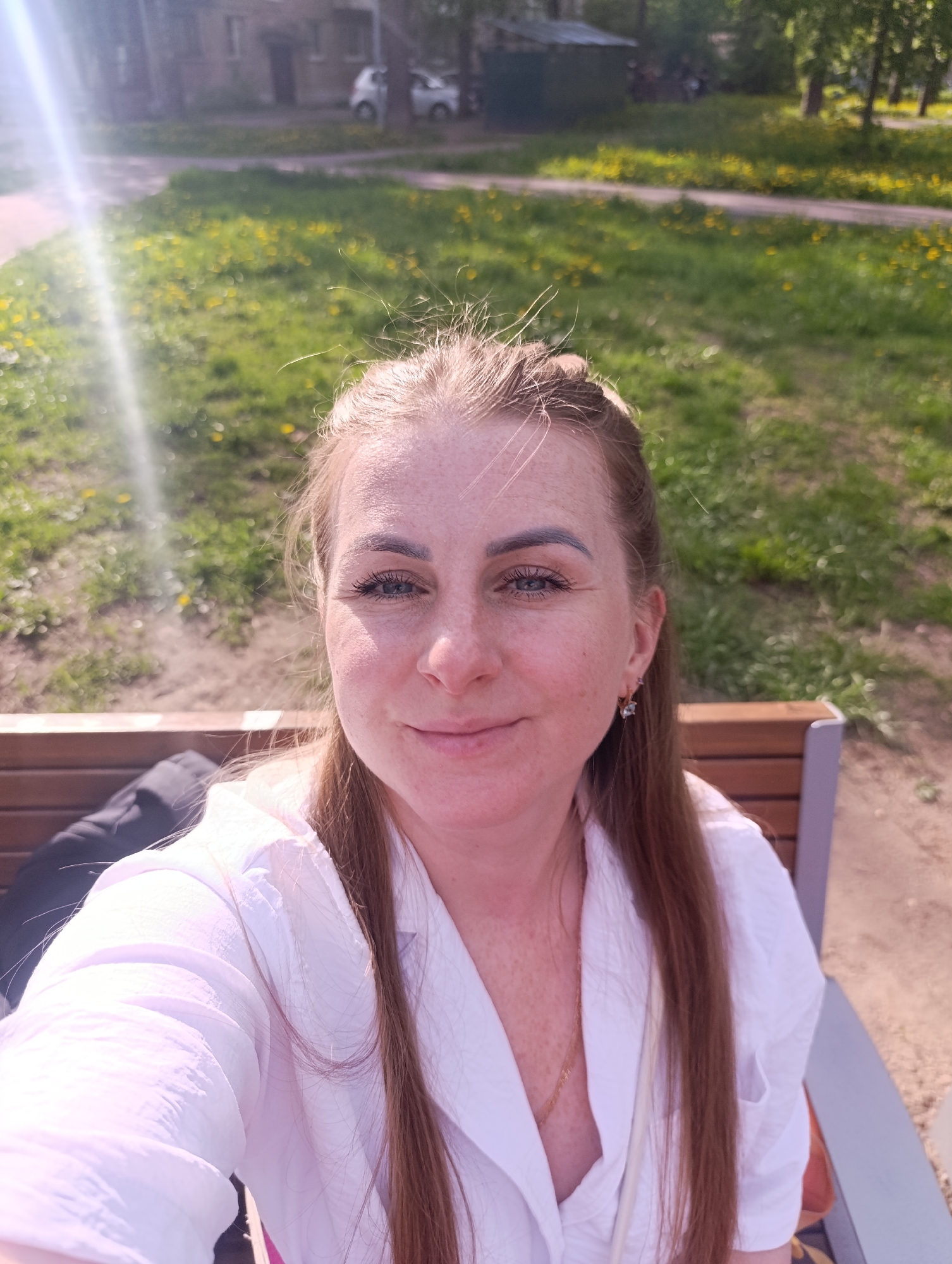 A woman is seated on a park bench in an outdoor setting, under bright sunlight. The location is within the city of Unknown, Unknown. The woman is wearing a white shirt. She has red hair and blue eyes. In the background is a grassy lawn and trees. A building and car are visible further in the background.