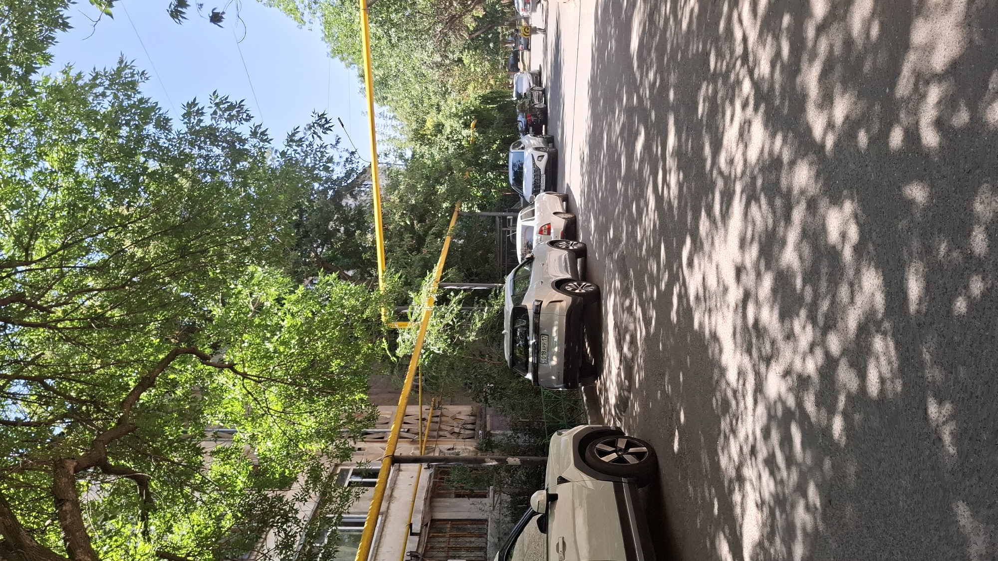 The image depicts a street scene in Shymkent, Kazakhstan, on a sunny day. Several cars are parked along the side of an asphalt road, which is dappled with sunlight filtering through the leaves of large trees. To the left, a multi-story apartment building is visible, with yellow pipes running along its facade. The building appears to be made of concrete or plaster and has windows with metal grates. The trees are deciduous, with full green foliage, and their branches extend over the road and building. In the background, more trees line the street, creating a shaded canopy. A few individuals are present, including a person walking away from the camera in the distance. The overall atmosphere is that of a residential urban environment during daytime.