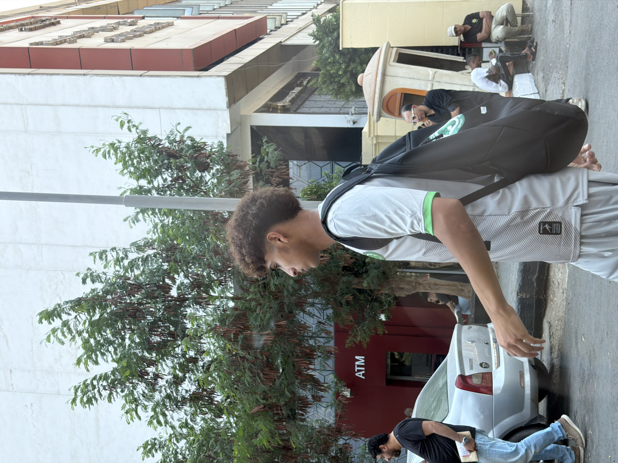 The image depicts a street scene in Cairo, Egypt, with a young person in the foreground walking from left to right. They are wearing a grey and white athletic shirt with green trim and white pants, and carrying a black backpack. To the left, a man in a black t-shirt and blue jeans walks towards the left, holding a book or paper. Behind them is a silver sedan. In the background, a building with a red lower section displaying an "ATM" sign is visible. Further in the background, there is a small, ornate booth-like structure and several other people, some seated. Trees with long, thin seed pods are present throughout the scene, and a grey lamppost stands near the center. The overall environment suggests a moderately busy urban street.
