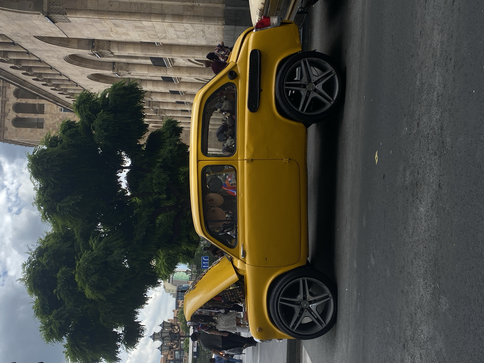 A vibrant street scene in Yerevan, Armenia, features a customized bright yellow classic car parked on the asphalt road. The car, possibly a modified Fiat 600 or similar microcar, is fitted with large, modern, five-spoke alloy wheels. Its front hood is propped open, revealing various items, seemingly hats or other merchandise, displayed inside. Several people are gathered near the open hood, looking at the items, and two individuals are visible inside the car through the rear window.
Behind the car, a large, lush green tree stands prominently, partially obscuring a grand, multi-story stone building. This building exhibits classical architecture with numerous arched windows and decorative elements. The setting suggests an urban environment, likely a city center. The sky is overcast with visible clouds, indicating a cloudy daytime. A blue road sign with white arrows is partially visible behind the car's open hood, and ornate streetlights can be seen further in the background to the right of the tree. Another vehicle is partially visible parked behind the yellow car, with its rear license plate partially readable, ending in "136". The scene captures a lively moment, possibly a street vendor displaying wares from the unique vehicle.
