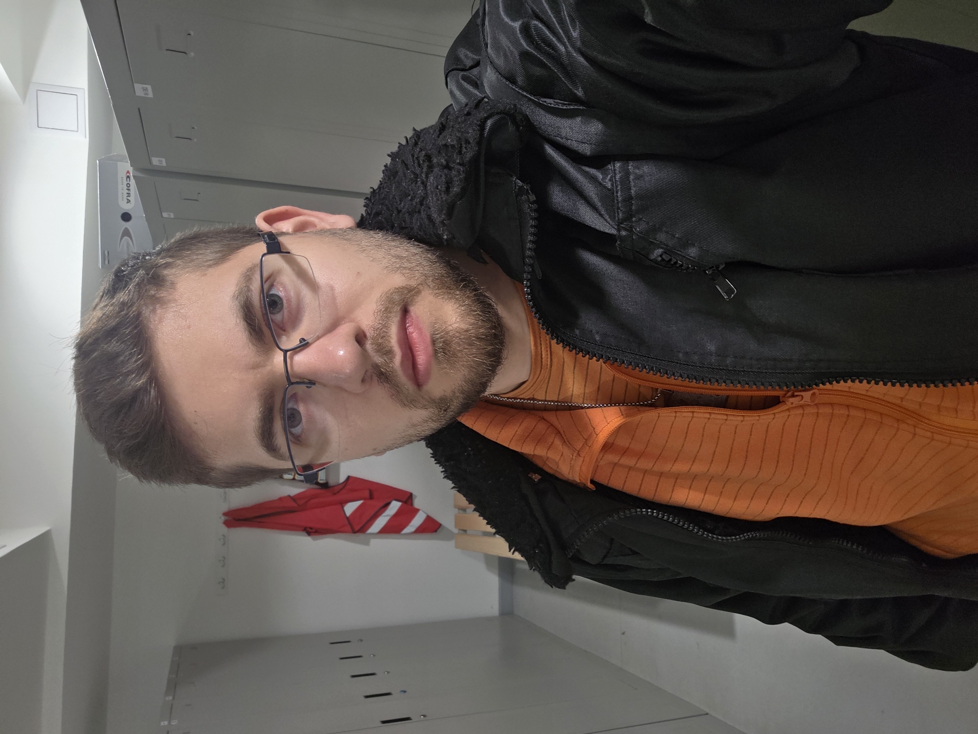 A man with short brown hair, a beard, and glasses is wearing an orange striped shirt and a black jacket. He appears to be in a changing room or storage area, with metal lockers and a red and white garment visible in the background. There is no information in the image to indicate the location is in Plovdiv, Bulgaria.
