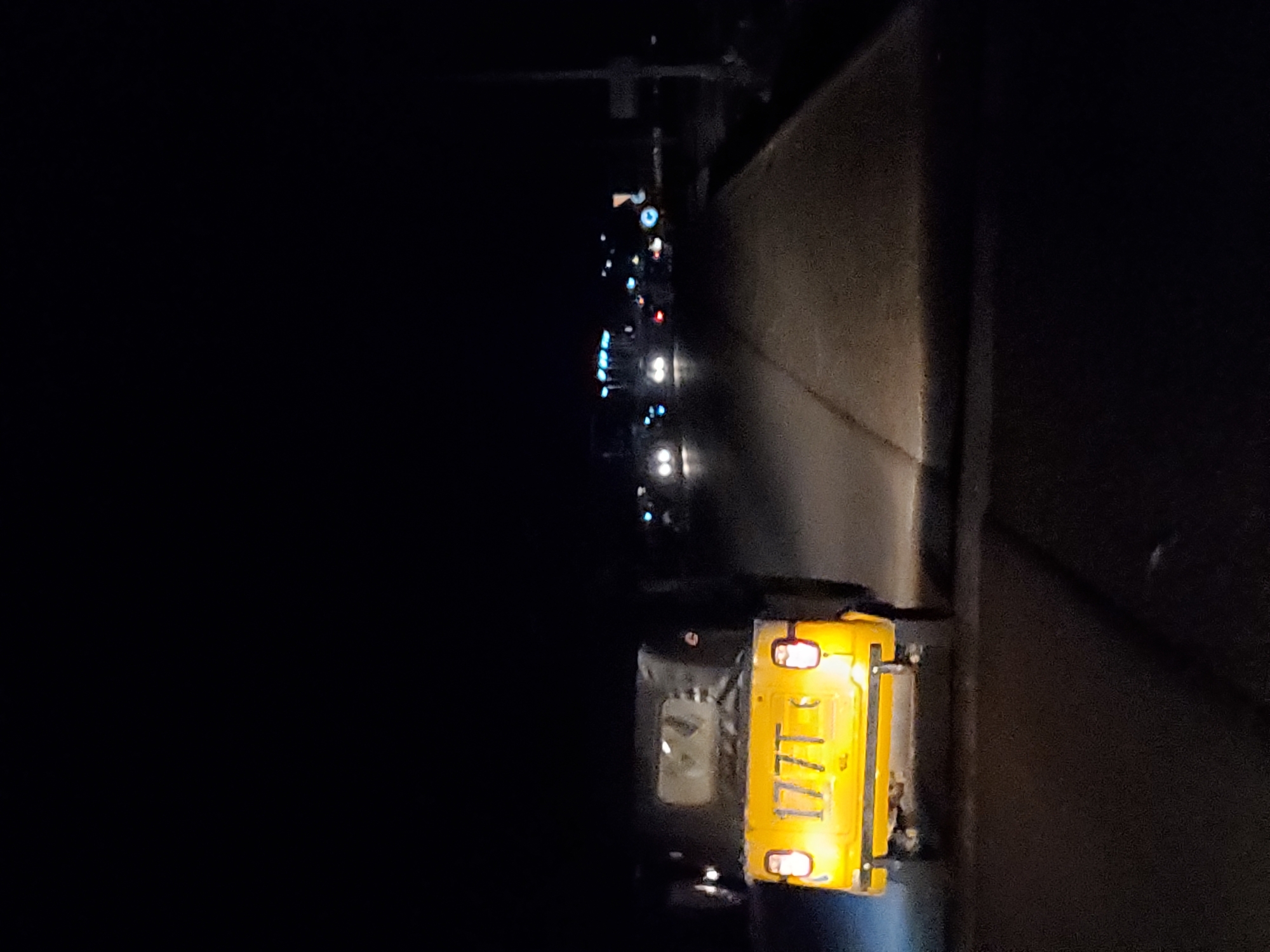 The image depicts a nighttime scene on a road in Jalingo, Nigeria. A yellow auto-rickshaw, identified by the license plate "177T," is prominently featured in the foreground, traveling away from the viewer. The vehicle's headlights and taillights are illuminated, casting a glow on the asphalt road. In the background, other vehicles with their lights on are visible, suggesting traffic or a gathering of vehicles. The environment appears to be a street or highway, with a dimly lit roadside and some distant lights that might indicate urban infrastructure or buildings. The overall ambiance is dark, characteristic of a nighttime urban setting.