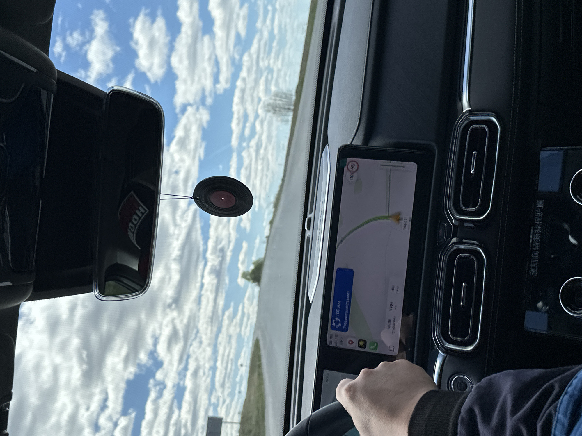 The image shows a view from inside a car, with the dashboard and parts of the steering wheel visible. The navigation system displays a map with a route indicating "1212 GM, Pulyanovskata" as the next direction, and the destination is "Doma 78 Km" with an estimated time of arrival at 11:00. In the rearview mirror, the reflections of a cloud-filled sky are visible, and a circular air freshener hangs from the mirror. The driver's hand is visible on the steering wheel.