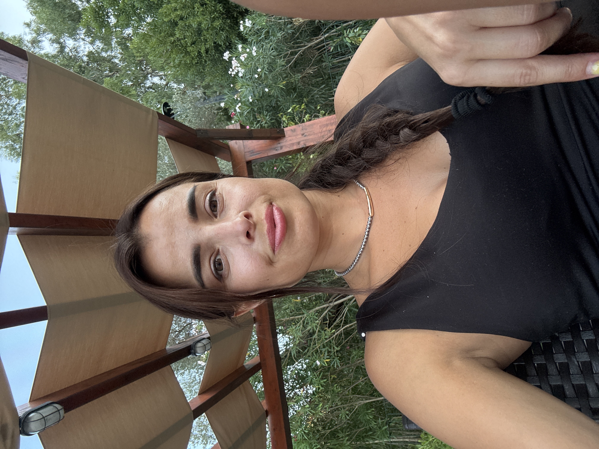 A woman with dark hair in a braid is posing for a selfie outdoors. She is wearing a black tank top and a silver necklace. She is seated under a wooden and canvas canopy, with lush green trees and white flowers in the background. The lighting suggests it is late afternoon or early evening.