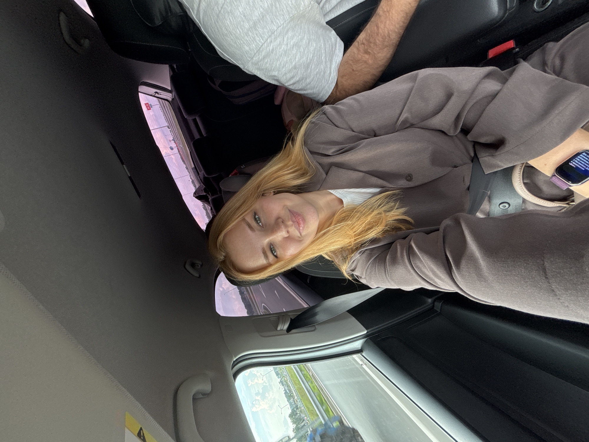 The image shows a young woman with blonde hair and fair skin, wearing a brown blazer over a white top, seated in the passenger seat of a car. She is smiling at the camera, which is held by an unseen person. Outside the car window, a highway with a blue tractor is visible, suggesting the car is in motion. The interior of the car features black upholstery and a grey headliner. A portion of another person's arm, possibly a male, wearing a grey t-shirt, is visible on the right side of the frame. The scene appears to be taking place in or around Kuznechikha, Russia, based on the provided location.
