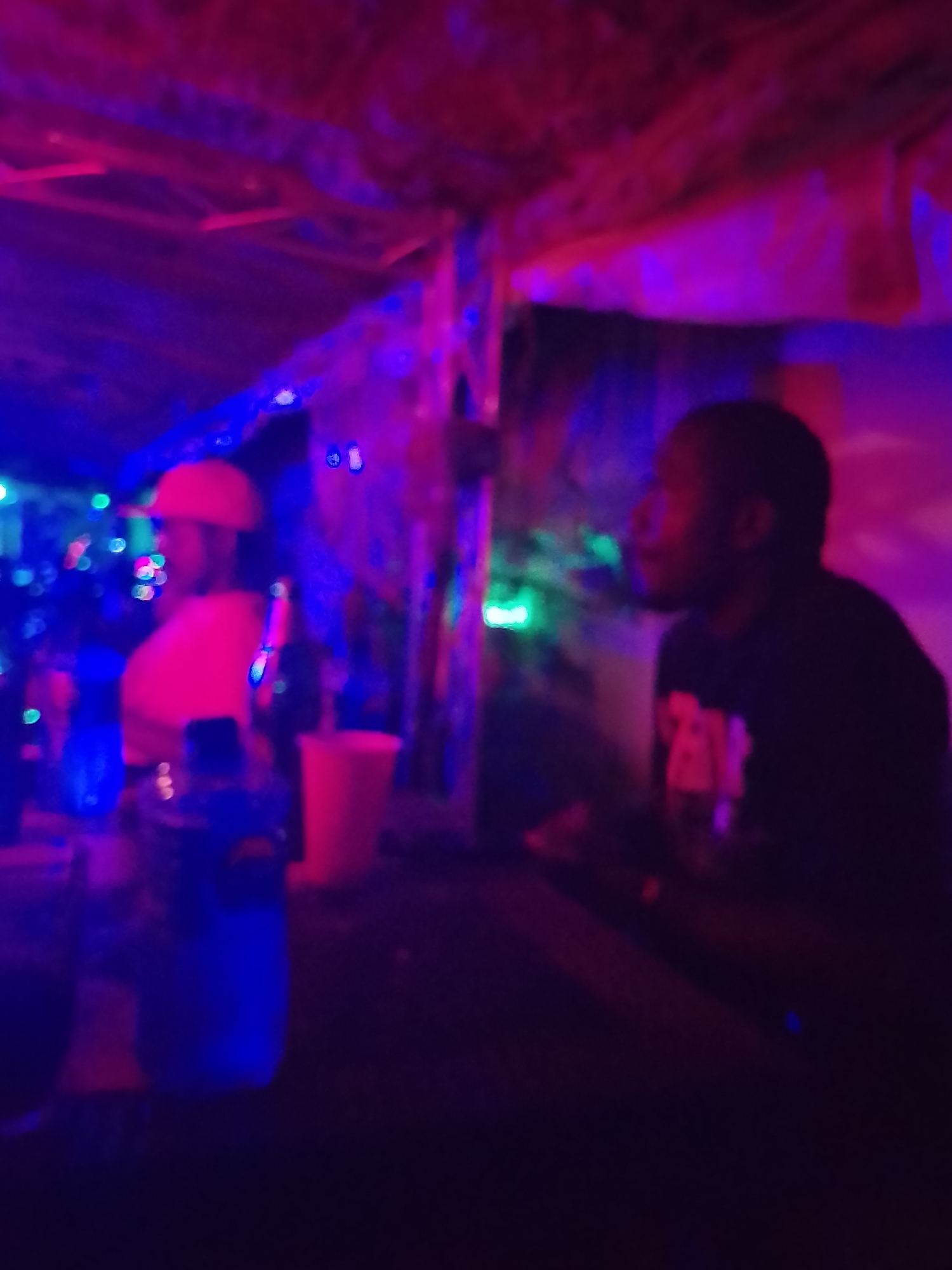 This image captures a vibrant, dimly lit indoor scene, likely a bar or club, bathed in strong artificial lighting dominated by purple, red, and blue hues. The overall aesthetic is blurred, suggesting movement or low light conditions.
Two individuals are visible, seated at what appears to be a bar or table. On the left, a person with dark skin is seen from the chest up, wearing a red cap or hat and a light-colored (possibly red or orange) shirt, facing towards the right. Their facial features are indistinct due to blur and lighting. Various bottles and cups are on the surface in front of them, including a prominent blue bottle and a red cup.
On the right, another person with dark skin is depicted in profile, looking towards the left. They are wearing a black t-shirt with a light-colored, blurry graphic or text on the chest. Their face is also softened by the image blur.
The background features numerous scattered light sources in blue, green, and red, creating bokeh effects that enhance the club-like atmosphere. The overhead structure appears to be made of rough, possibly rustic materials like wood or thatch. The scene depicts a social gathering or an evening out in Yelwa, Nigeria, characterized by its lively and colorful artificial lighting. No clear text is visible in the image.