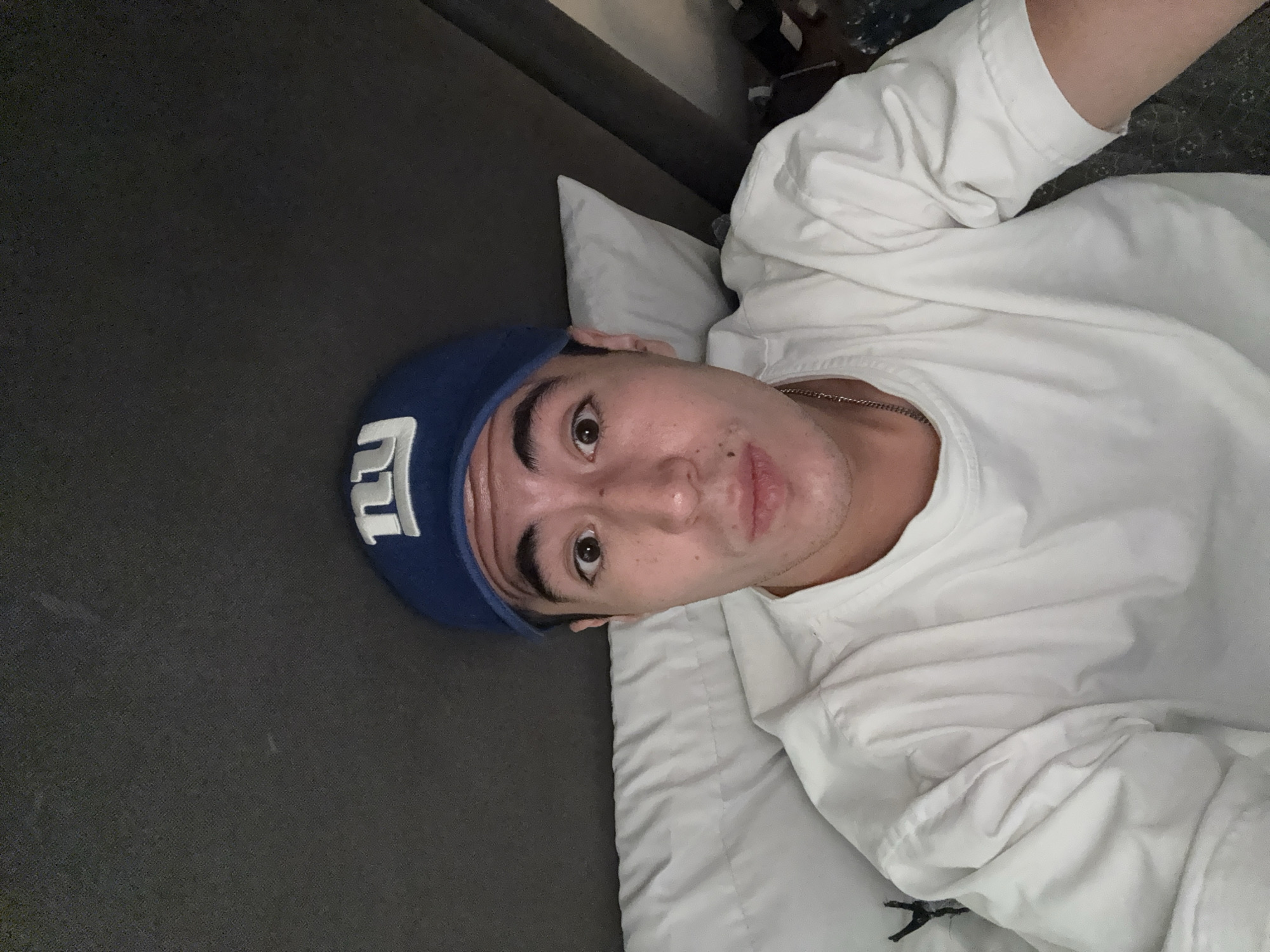 A man lies on a grey couch, propped up by a white pillow. He is wearing a white long-sleeved shirt and a blue baseball cap with a white "NY" logo. A silver chain is visible around his neck. The scene is likely indoors, but the location is not San Bernardino Tlaxcalancingo, Mexico, based on the visual information.
