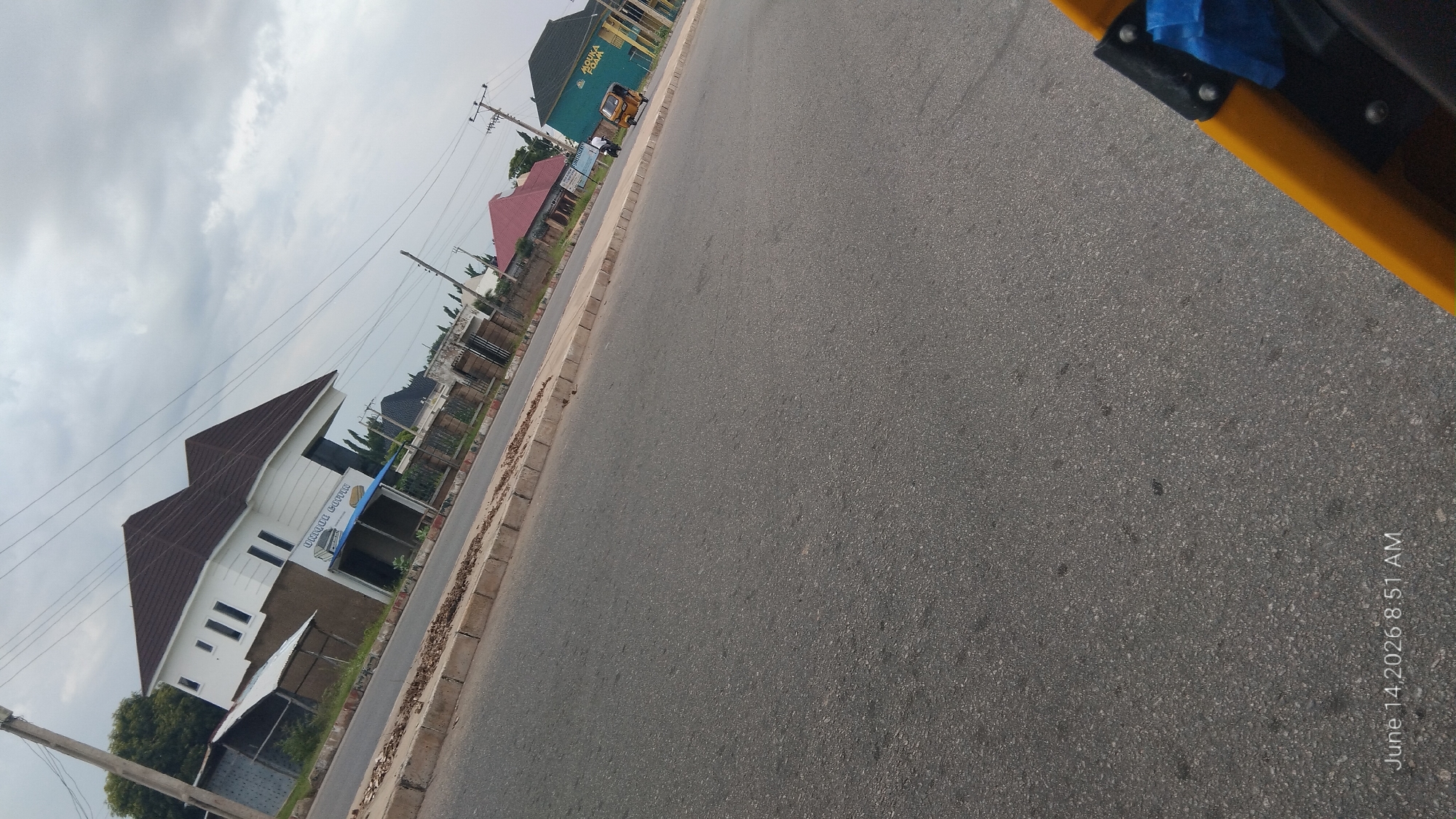 The image shows a street scene in Jalingo, Nigeria. A two-story building with a white facade and a brown roof is visible on the left side of the road, with signage that reads "Unique Cabin". Further down the street, a turquoise building with "MOUKA FOAM" written on it is on the right. A yellow three-wheeled vehicle is parked in front of this building. The sky is overcast with clouds, and the road appears to be asphalt. Power lines are strung overhead. The timestamp "June 14, 2026 8:51 AM" is visible in the lower-left corner. A portion of a yellow object, possibly part of a vehicle or structure, is visible in the foreground.