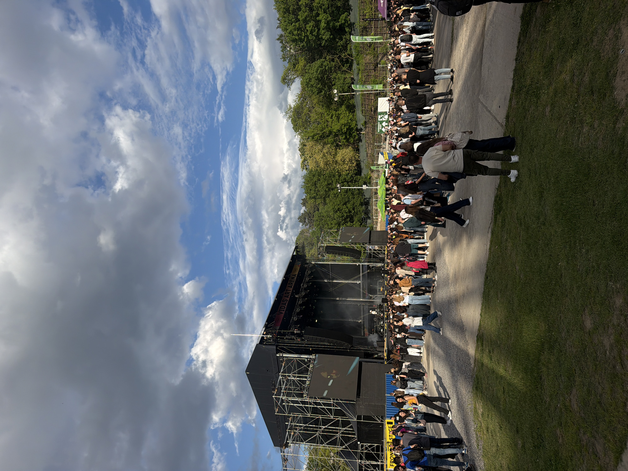 A crowd of people is gathered in front of an outdoor stage, likely at a concert or festival. The stage is a large structure with scaffolding and screens. Some people are standing, while others are walking or interacting. The sky is partially cloudy with patches of blue. The location is likely Vitosha mountain in Sofia, Bulgaria.