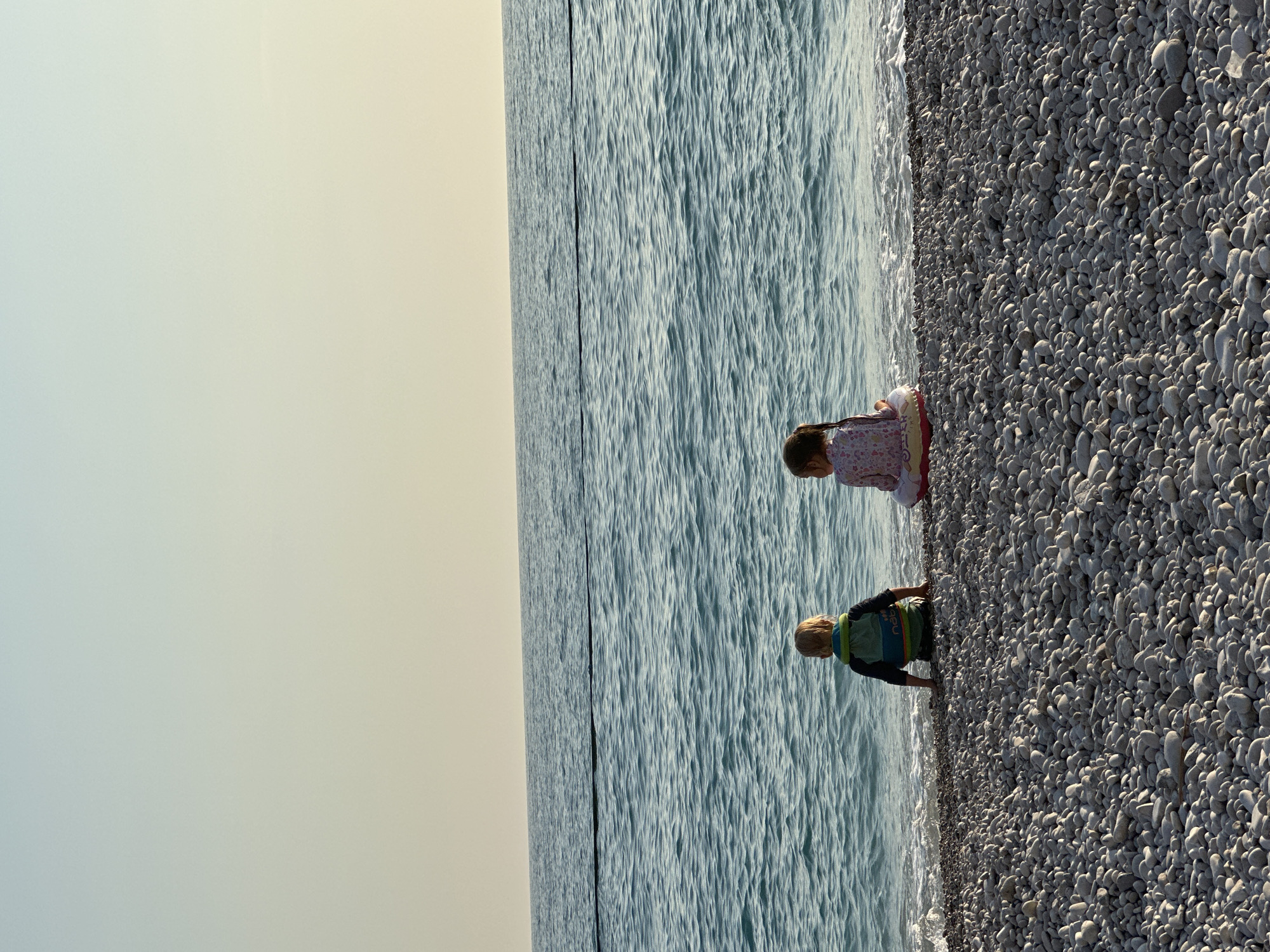 The scene depicts two young children observed from behind, seated on a pebble beach in Fethiye, Turkey, facing the Mediterranean Sea. The time of day is late afternoon or early evening, indicated by the soft gradient of the sky from pale blue overhead to light yellow at the horizon. The child on the left, a boy with blonde hair, wears a dark long-sleeved swim shirt and a green and blue buoyancy aid with "Nabaiji" visible on the back, sitting directly on the greyish-white pebbles at the water's edge. The child on the right, a girl with dark hair tied in a ponytail, wears a light-colored, patterned long-sleeved swim shirt and sits on a pink, white, and yellow inflatable ring, also at the water's edge. Gentle waves lap the shore in front of them. A thin, dark line, possibly a buoy rope or demarcation, extends horizontally across the blue-green sea in the mid-distance. The foreground of the image consists entirely of uniform, smooth, light-colored pebbles.