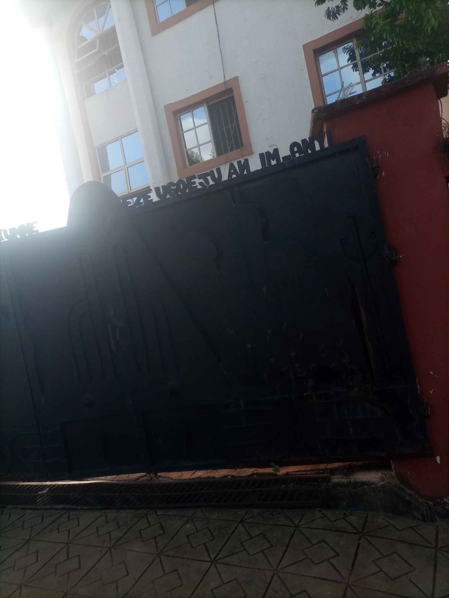 The image shows a dark, ornate metal gate in front of a white building with arched and rectangular windows. Above the gate, a sign with black lettering is partially visible and reads "EZE UGOESTU AM IM ANY". The building appears to be multi-story and features orange trim around the windows. To the right of the gate and building, a red wall is visible. The ground in front of the gate is paved with patterned tiles. Sunlight is strongly entering the frame from the left, creating a bright lens flare and obscuring some details of the building and gate. The scene is likely outdoors in a city.