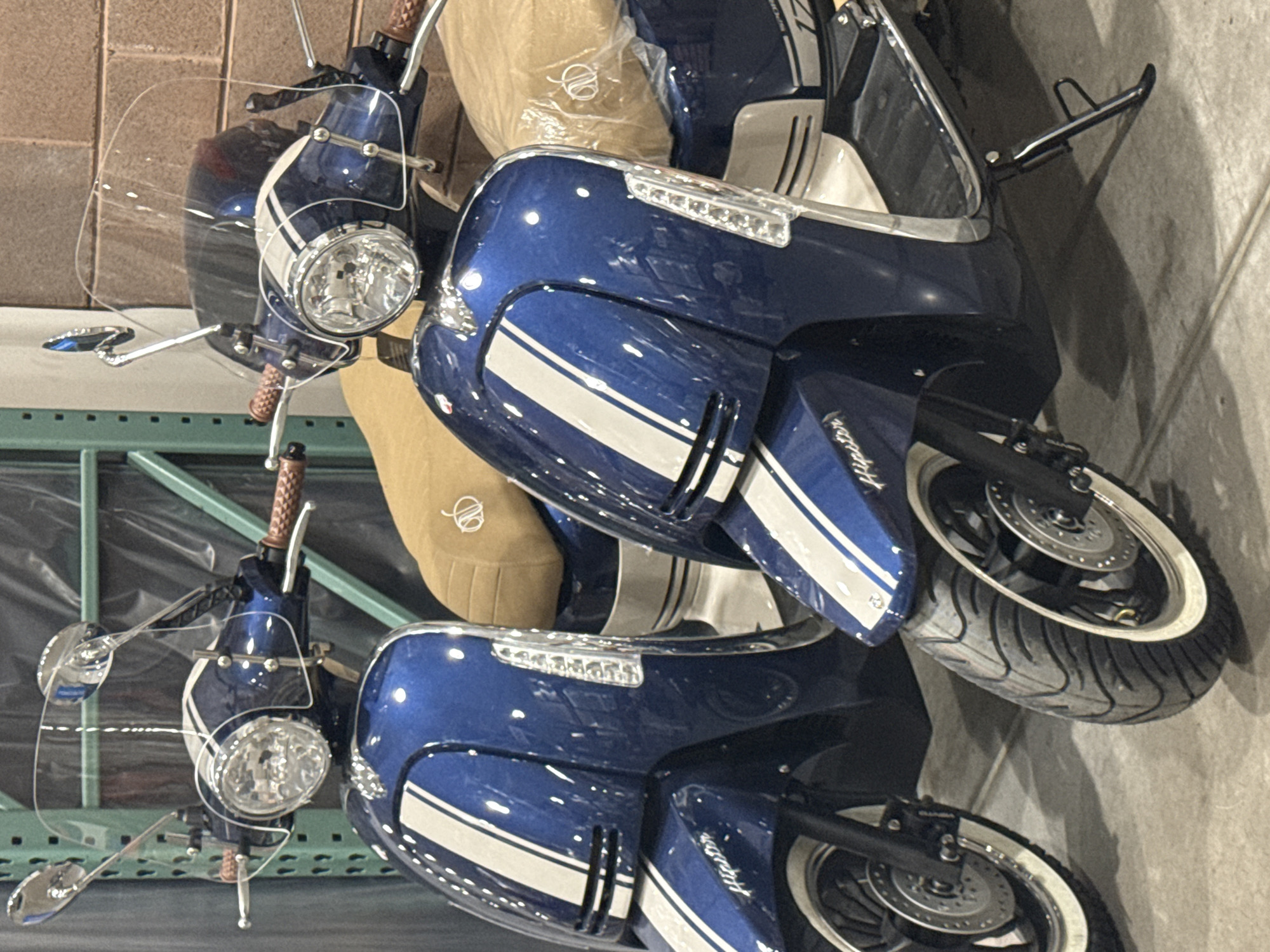 The image shows two blue scooters with white racing stripes parked indoors. Each scooter has a windshield, a tan seat, and white-walled tires. The location is likely a warehouse or storage facility, as suggested by the concrete floor and metal racking in the background. No people are visible. The scooters appear to be new or well-maintained. The "Hipster" logo is visible on the front fender of both scooters. There is no indication that the image was taken in Puebla, Mexico.