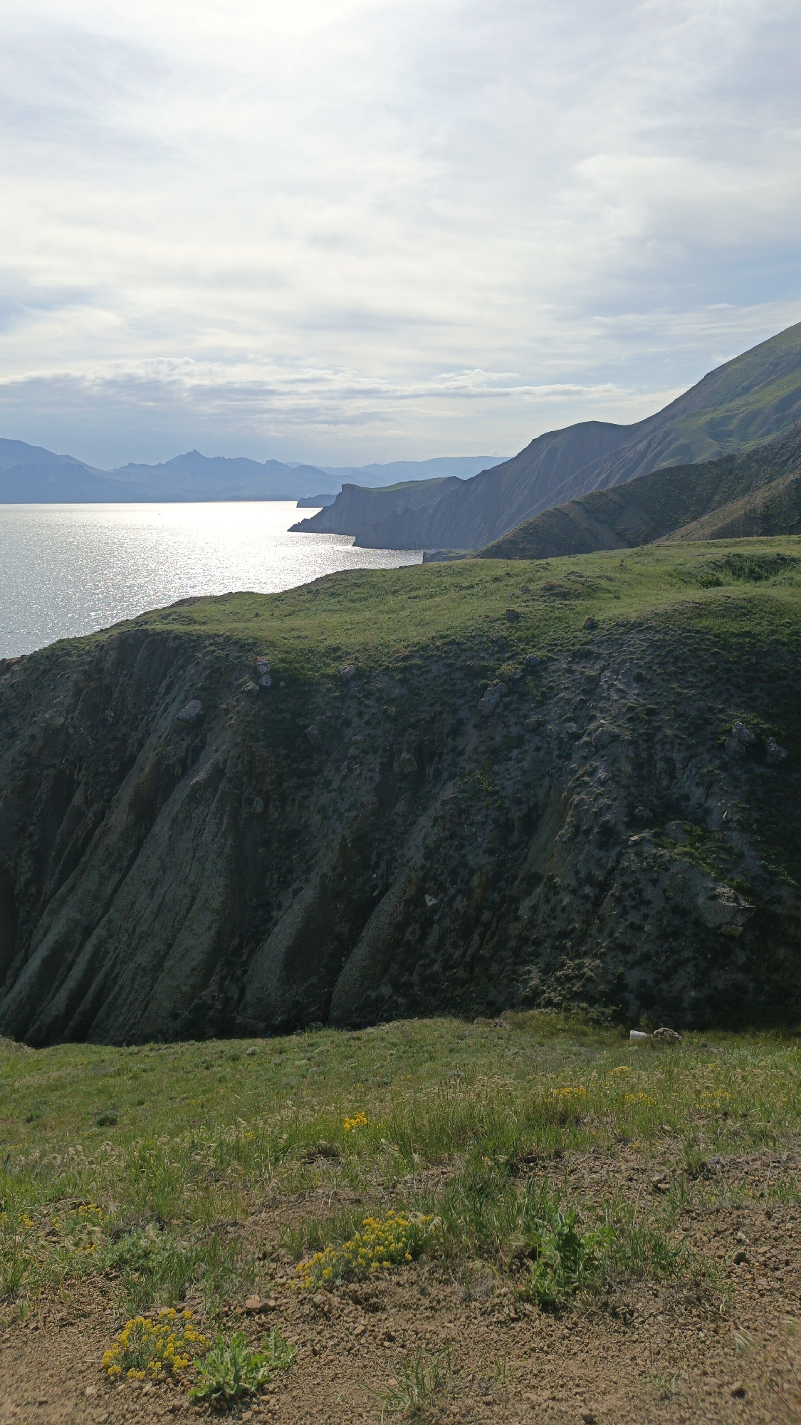 The image captures a coastal landscape. A sea or large lake occupies the left side of the frame, reflecting sunlight. Adjacent to the water is a steep, grassy cliffside. The landscape includes rolling hills, the slopes covered in vegetation. In the foreground, low-lying vegetation including small yellow flowers grow. The sky is overcast with a mix of clouds. The location is Unknown, Unknown.