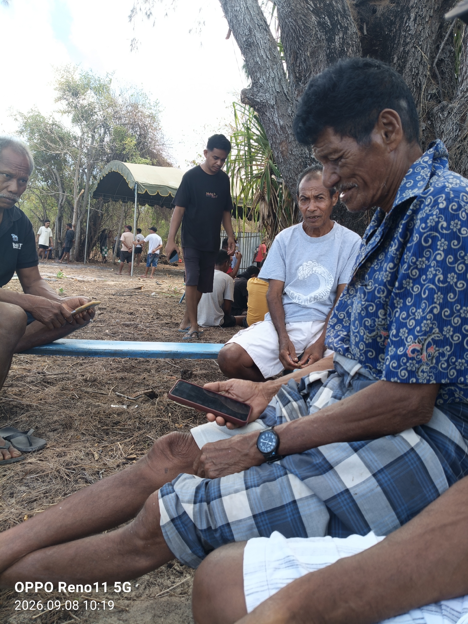 In an outdoor setting, possibly a beach or rural area, several individuals are gathered. In the foreground, a man wearing a blue and white patterned shirt and plaid shorts sits on the ground. He is holding a dark-colored smartphone in his right hand and has a watch on his left wrist. To his left, another man in a gray t-shirt and white shorts sits with his hands in his lap. To the left of these two men, a third man in a black t-shirt with a logo is partially visible, holding a light-colored object. In the background, a large tree with rough bark is prominent. Behind the people, there is a tent with a green and yellow canopy. Other individuals are seen in the middle ground and background, some standing and some sitting, with trees and dry vegetation surrounding them. The ground is covered with dry leaves and debris. A timestamp in the lower left corner indicates "OPPO Reno11 5G 2026.09.08 10:19".