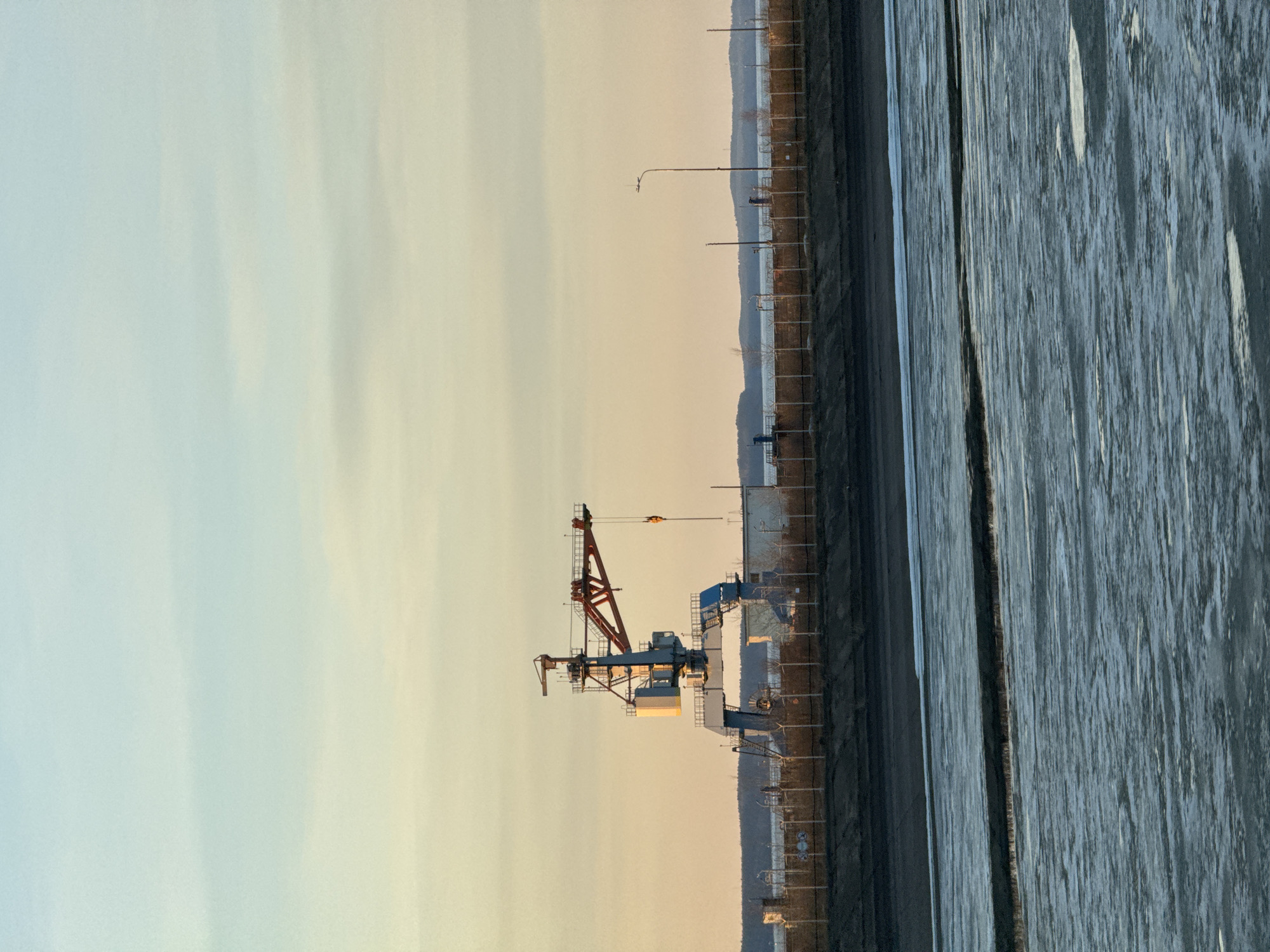 The image captures an industrial waterscape at what appears to be either sunrise or sunset, given the gradient of soft light in the sky transitioning from warm yellow-orange hues near the horizon to a pale, cool blue overhead. The scene is devoid of people.
In the foreground and mid-ground, a large body of water occupies the right side of the frame. Its surface is textured, possibly indicating ice cover, choppy water, or a reflection pattern, appearing in shades of grey and blue. Adjacent to the water, running horizontally across the image, is a dark strip of land or shoreline.
On the left side of this land strip, a prominent industrial crane stands. The crane is painted in contrasting red and grey-blue colors, featuring a complex structure of beams, platforms, and a visible hoist mechanism with a yellow hook. Further along the shoreline, behind the crane, are several tall utility poles or streetlights, and a fence line can be discerned. In the background, a series of low, rolling hills or mountains form a distant horizon, appearing as dark silhouettes against the bright sky.
No specific activity is taking place, and the scene presents a quiet, possibly cold, environment. Given the context of Chaikovsky, Russia, this could be a view of the Kama River or Votkinsk Reservoir, potentially an industrial port area. There is no visible text in the image.