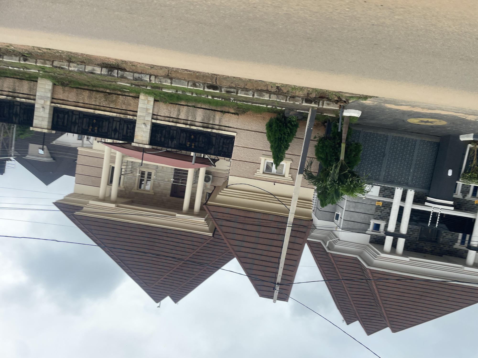 The image captures a street view in Abuja, Nigeria, showcasing a row of modern, multi-story houses. The architecture is characterized by prominent peaked brown roofs with a tiled texture, large white columns supporting overhangs, and a combination of light beige and grey facade materials. The houses are enclosed by decorative black metal gates and stone-clad walls. In the foreground, a paved road runs horizontally, with a grassy verge and a low curb separating it from the properties. The sky is overcast with grey clouds. The scene is devoid of people or visible vehicles, presenting a static, residential environment. Power lines are visible crisscrossing the sky.
