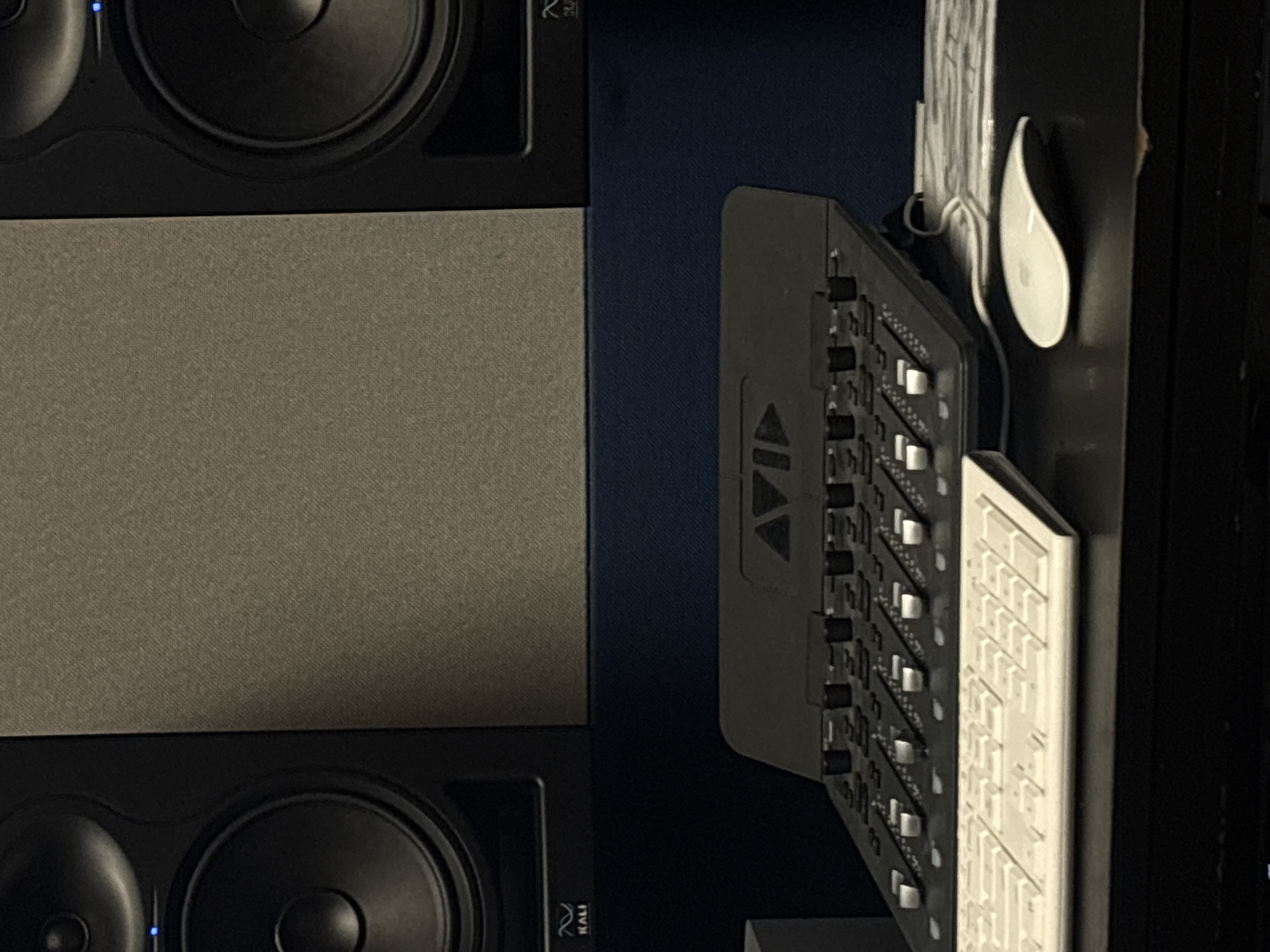 The image shows a close-up, slightly rotated view of a desk setup. On the left side are two Kali Audio studio monitors positioned vertically, with a rectangular soundproofing panel between them. To the right, an Avid mixing console is placed alongside a white Apple Magic Keyboard and Magic Mouse. The environment appears to be a home studio or workspace, with dark blue walls in the background. There are no people visible in the image. There is no evidence to determine the location of the image.