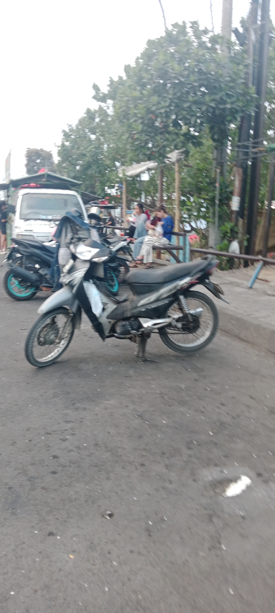 In Ngemplak, Indonesia, a scene unfolds on an asphalt road with a slight incline. Two motorcycles are prominent: a silver one in the foreground with its kickstand down, and a black one slightly behind it with bright teal rims. Behind the motorcycles, a white van is parked, suggesting a commercial or service vehicle. In the background, a group of people are gathered, some seated and interacting. Lush green trees provide a backdrop to the scene, with some foliage suggesting a tropical environment. A wooden fence and a utility pole are also visible elements of the surroundings.
