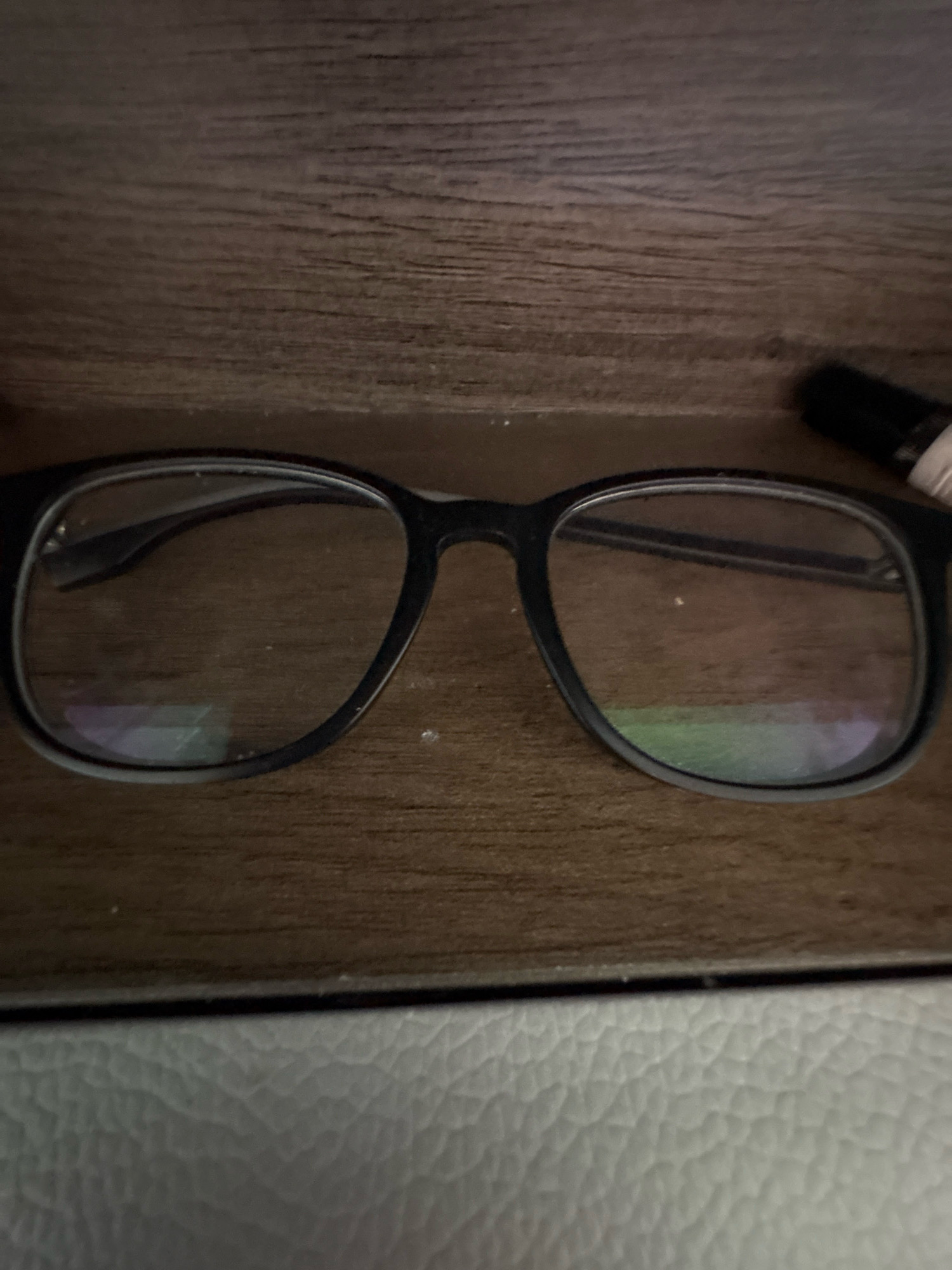 The image shows a pair of black-rimmed glasses resting inside a wooden glasses case. The case has a grainy, wood-like texture. The glasses are positioned horizontally, with the lenses facing upwards. The lenses are clear and appear to have some light reflections on them. To the right of the glasses, a black object with a white tip is partially visible, possibly a pen or a marker. The bottom of the image reveals a textured white surface, likely the exterior of the glasses case or a surrounding surface. The lighting in the image is dim, creating shadows and highlighting the textures of the objects. No people or specific locations are depicted.