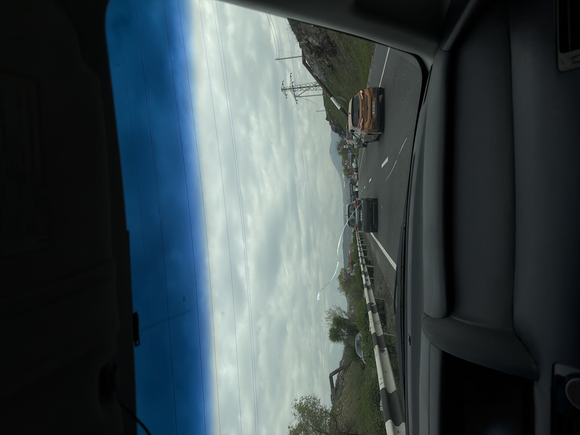 Captured from inside a vehicle, the image presents a view of a road scene in Arzni, Armenia. The road shows light traffic consisting of cars, with some appearing to be stopped in the roadway. The sky is overcast, creating a dull background over the scene. Infrastructure along the roadside includes utility poles.