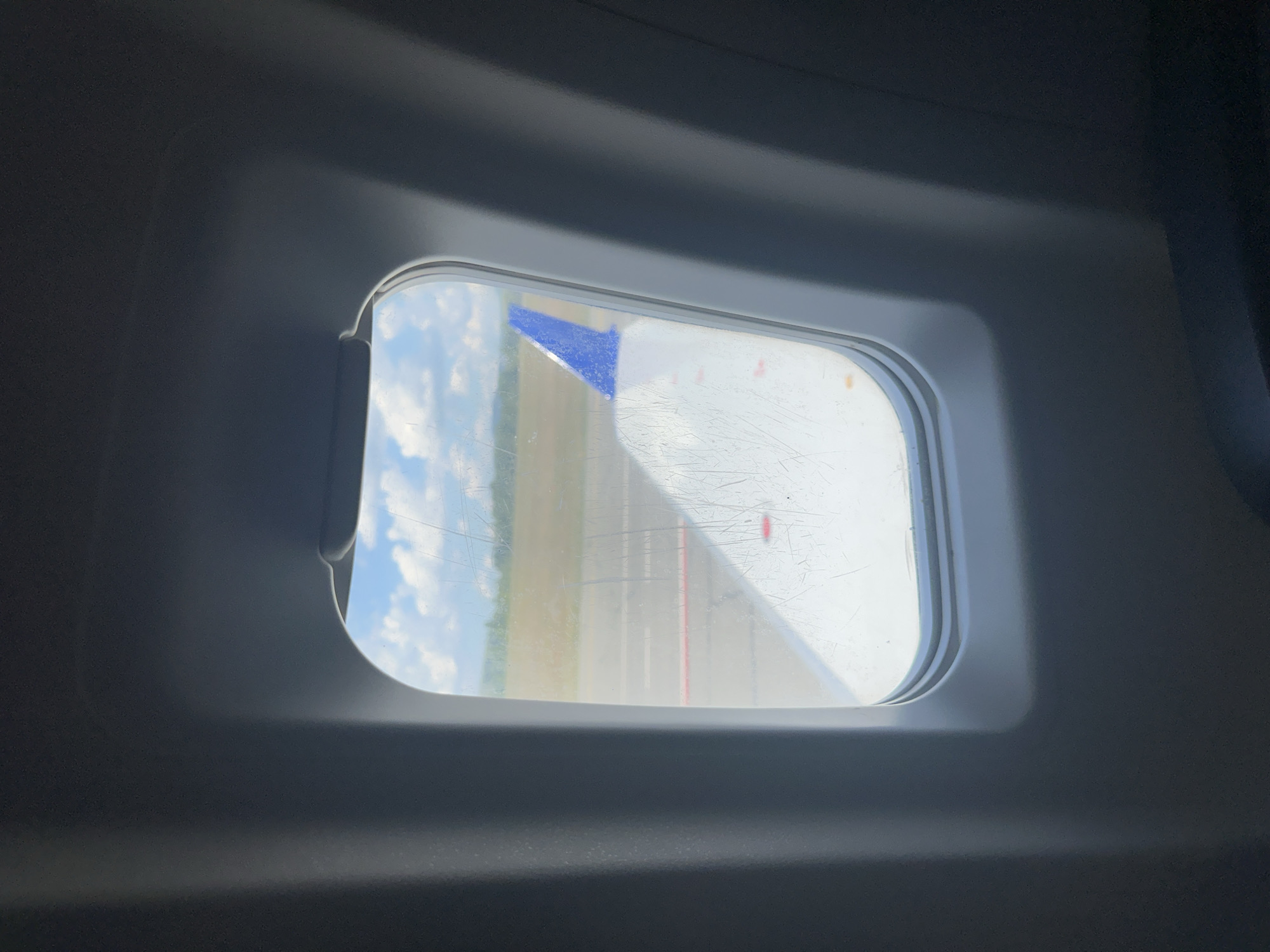 An interior view from an airplane window shows a wing with a blue tip, likely during takeoff or landing. The window is smudged with numerous scratches. Outside, a runway is visible under a cloudy blue sky, with a line of trees in the distance. The scene is devoid of any recognizable landmarks or features that could indicate the specific location within Maima, Russia.