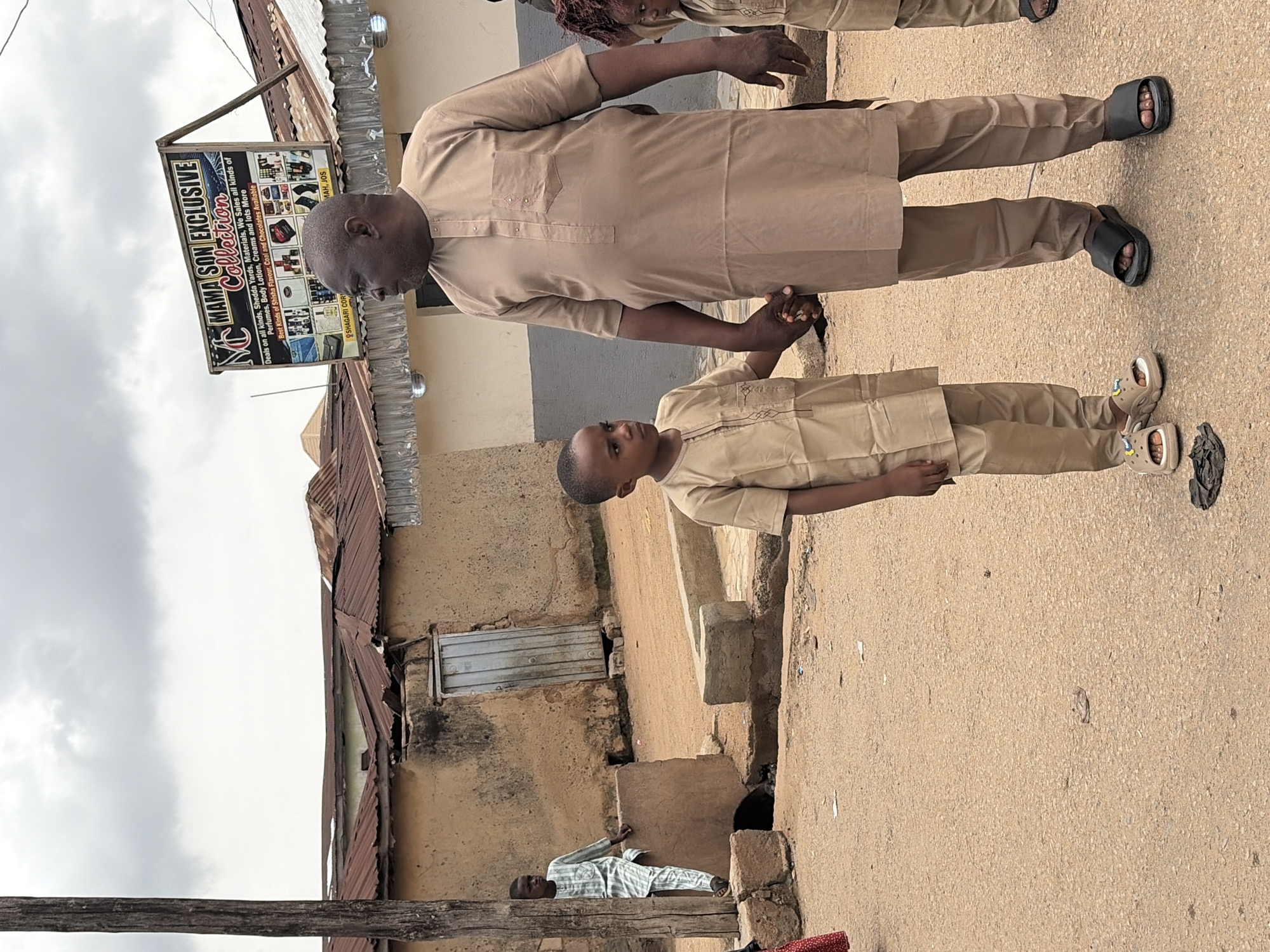 The image depicts a street scene in Rafin Par, Nigeria. In the foreground, a man and a young boy are holding hands and standing on a dirt road. Both are wearing traditional Nigerian attire. The man is in a tan, long-sleeved tunic and trousers. The boy is in a matching tan tunic and trousers. Behind them, the environment consists of simple, unpainted buildings with corrugated metal roofs. To the left, another boy in a striped tunic and trousers stands near a wooden post. A sign for "Mama Son Exclusive Collection" is visible on one of the buildings, advertising various goods. The sky is overcast. The overall impression is of a typical day in a residential area of a Nigerian town.