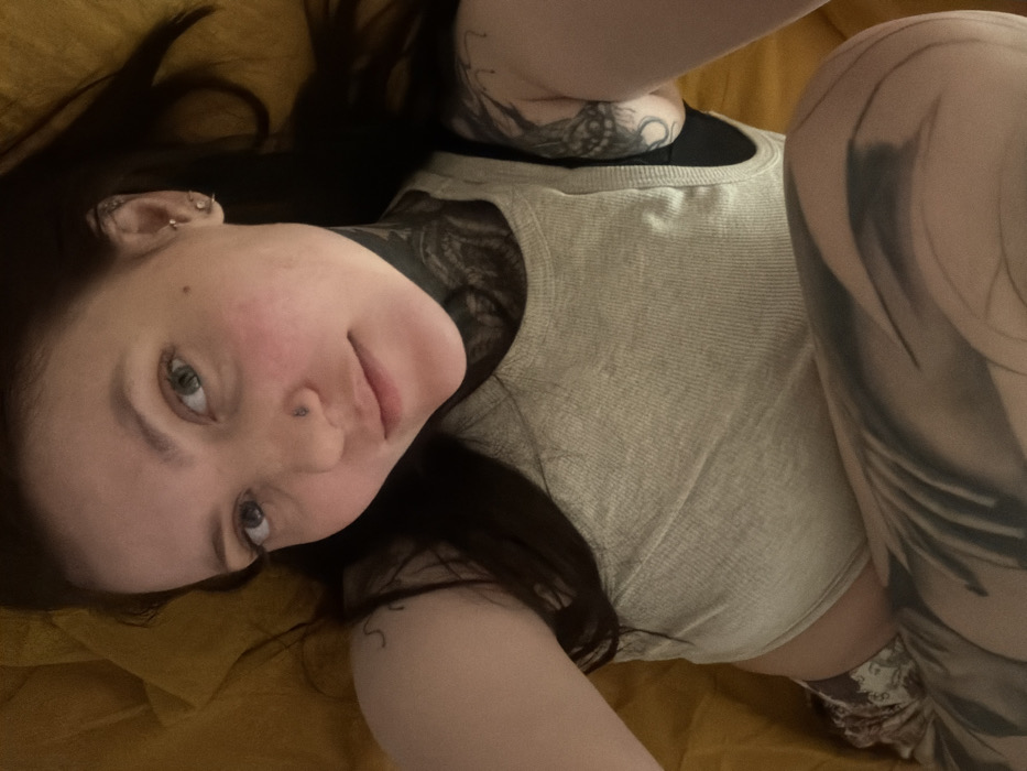 A woman with long dark hair and fair skin, adorned with numerous visible tattoos, rests on a mustard-colored surface. She has a nose piercing and multiple ear piercings. Her expression is gentle as she looks at the camera. She wears a light-colored tank top over a black undergarment. The visible tattoos include prominent designs on her neck, arm, and leg. The overall lighting is soft and diffuse, creating a casual and intimate feel.