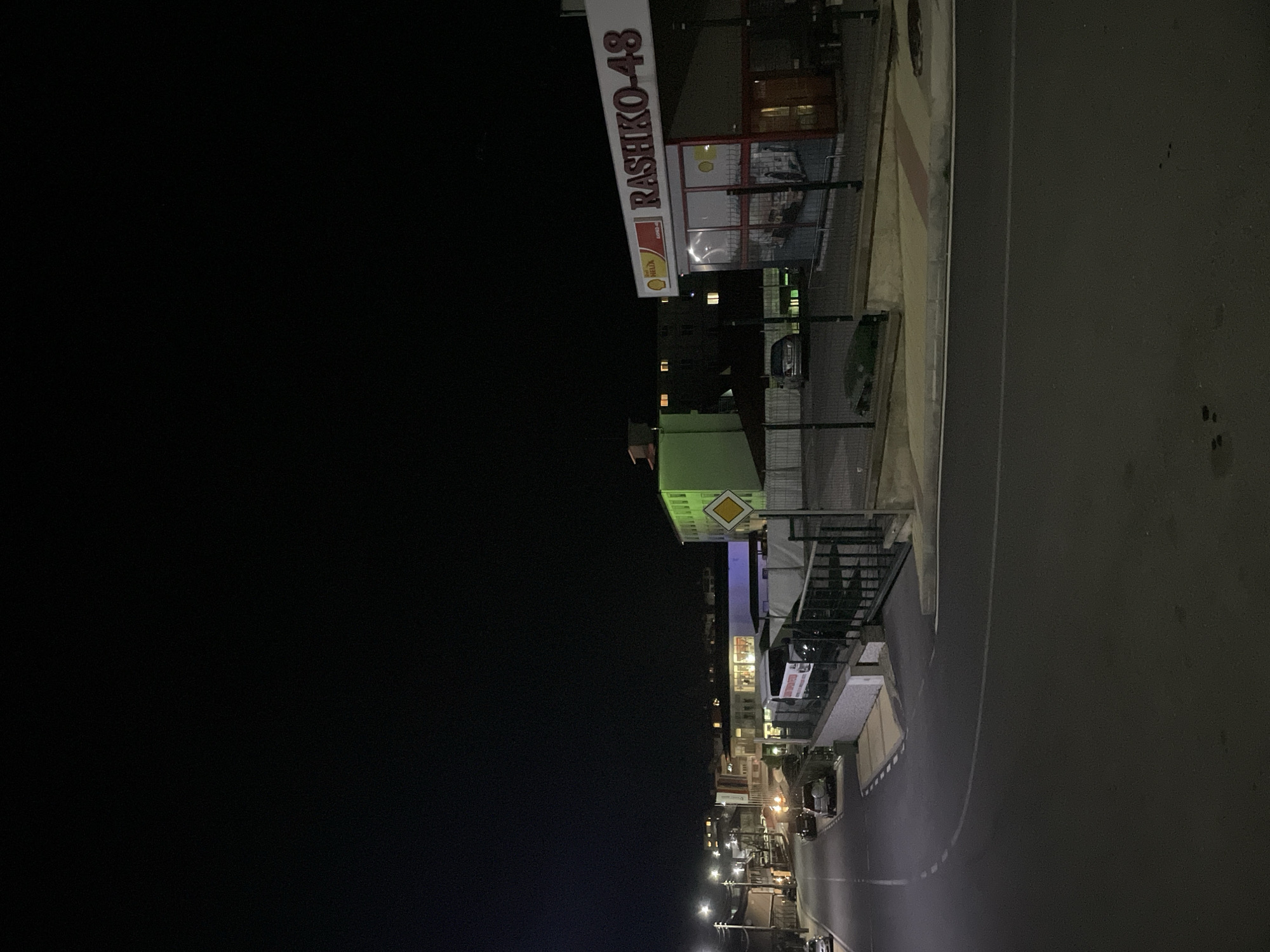 This image shows a night scene in Zlatograd, Bulgaria. The view is looking down a street with street lights illuminating the road. The “RASHKO-48” Shell gas station is visible in the upper right corner of the image, a building with the lights on. Several buildings are aligned along the street with multiple street lights illuminating the street. A road is present to the right. The sky is dark black to the left of the frame.