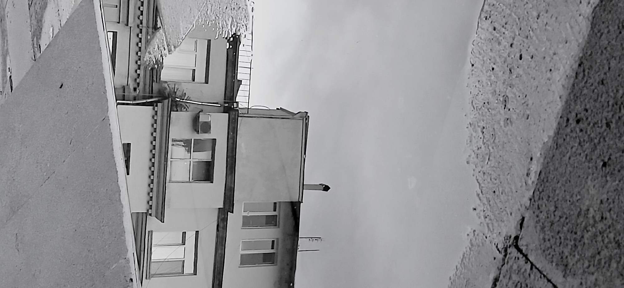 The image depicts a reflection of a building in a body of water, possibly a puddle or a small pond, against a grey sky. The reflection is upside down relative to the building. The building appears to be two stories high and is painted white with a dark roof. It has several windows, some with dark frames. There is also an air conditioning unit visible on the side of the building. The foreground of the image shows a textured surface, possibly concrete or asphalt, with some scattered debris. The scene is devoid of any people or visible activity. The image is a black and white photograph with a slightly grainy texture. The orientation of the photograph suggests it was taken with the camera pointing downwards towards the reflective surface.