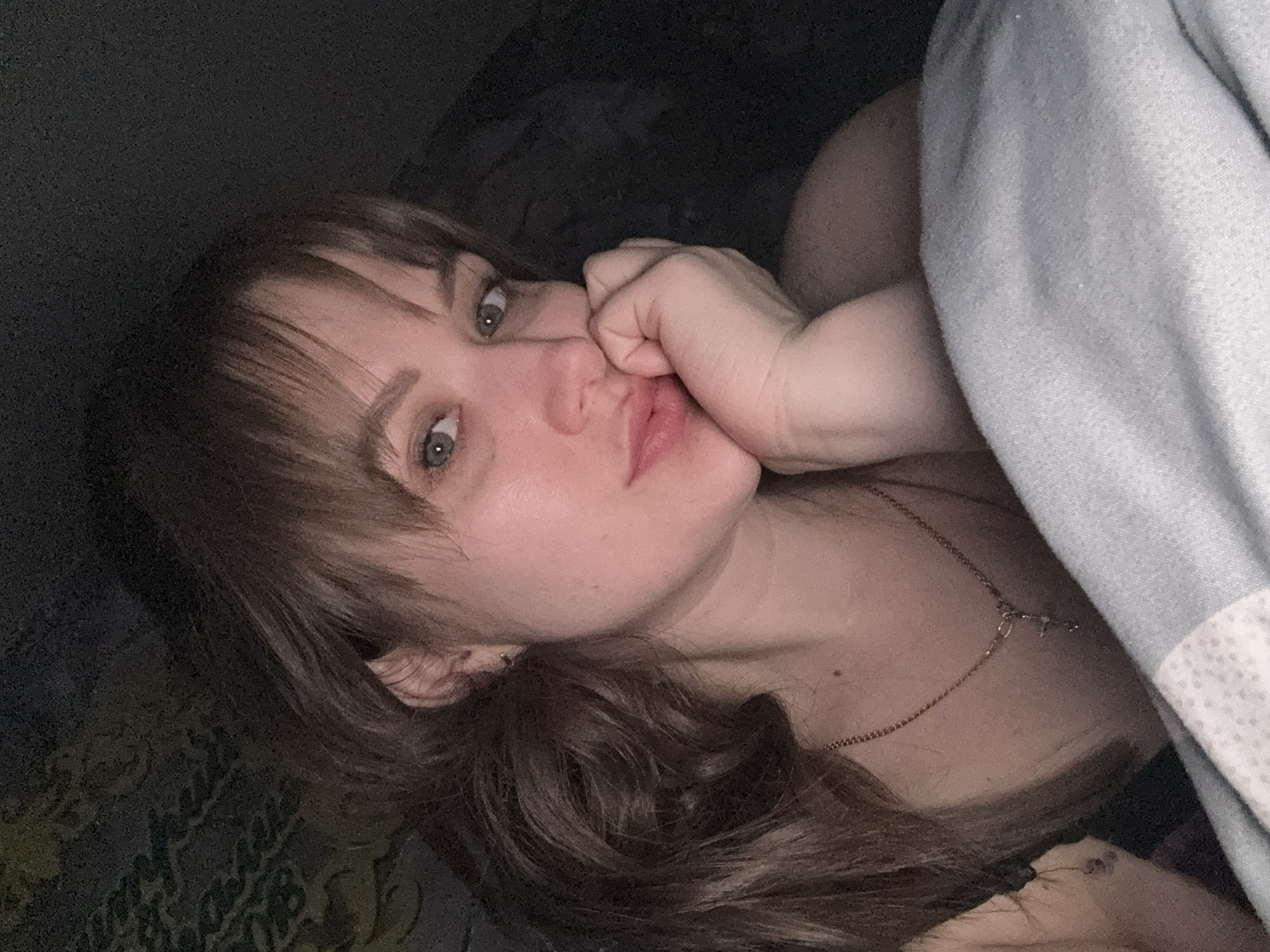 A woman lies in bed. She has brown hair with bangs and is wearing a gold necklace. She is holding her hand near her face. A light gray blanket is near her right side. The environment appears to be a dark bedroom. It is not possible to confirm the location as Chaikovsky, Russia.
