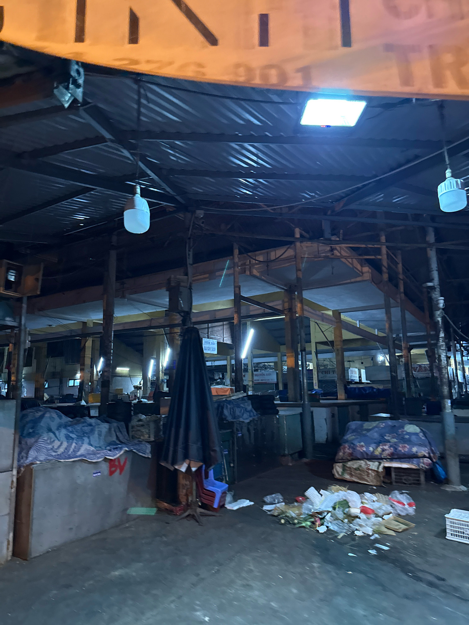 The image shows a nighttime view of a market stall in Nha Trang, Vietnam. The stall appears to be in a state of disrepair or is closed for the night. An orange banner with some lettering is visible at the top of the image, partially obscuring the corrugated metal roof of the market. Several fluorescent lights are on, illuminating the interior. A large, dark umbrella is set up in the foreground, with a small blue plastic chair stacked on top of others nearby. In front of the umbrella, a pile of garbage, including plastic bags, food scraps, and paper, is scattered on the concrete floor. To the left, a concrete counter is covered with a blue and white patterned tarp. Further back, other market stalls with tarps and various items are visible. The overall impression is one of a temporarily abandoned or neglected market setting.