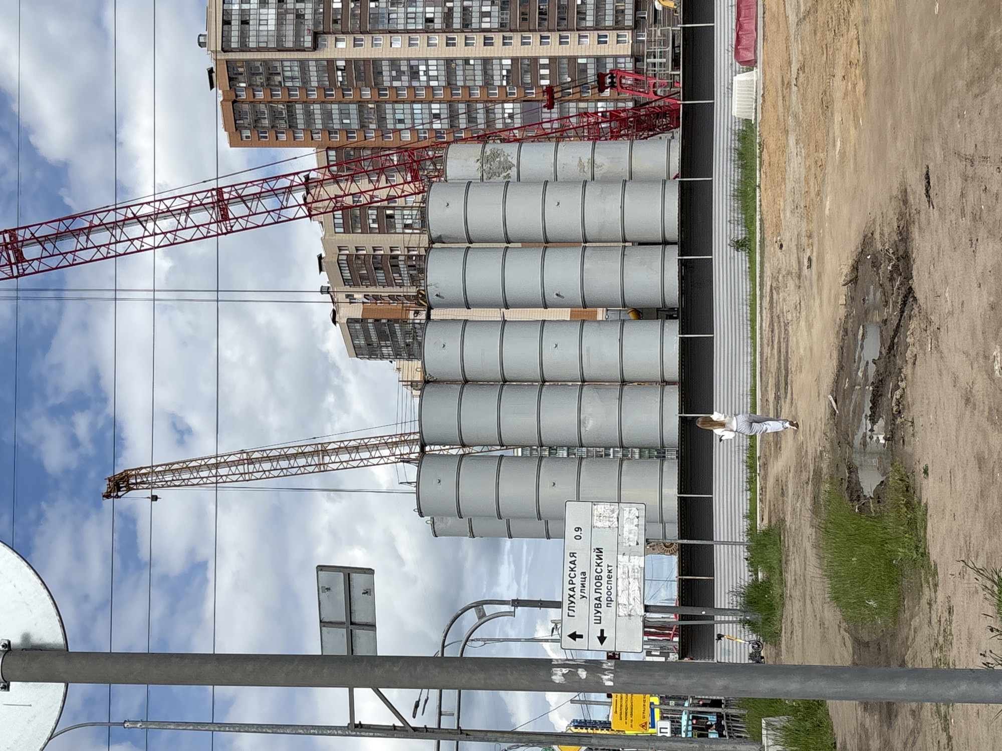 The image captures an urban construction scene under a partially cloudy sky in Saint Petersburg, Russia. The perspective suggests a view from a street or sidewalk overlooking a development area.
In the midground, a dirt ground covers most of the visible area, interspersed with patches of grass and several muddy puddles. A person, appearing to be a woman with long hair, dressed in a white top and light-colored striped pants, walks away from the camera on the dirt path. To her left, dominating the middle of the frame, are several large, grey, cylindrical silos or storage tanks, stacked horizontally, likely containing materials for the construction. A yellow electric scooter lies on the dirt nearby.
The background is filled with evidence of construction and urban development. Two large tower cranes are prominent against the sky: one is red and extends across the upper-mid section, and another, yellow and grey, is visible further to the left. Behind the silos and cranes, several tall, multi-story residential buildings, likely under construction or newly completed, stand with numerous windows.
In the foreground, on the right side, a white rectangular street sign with black text and arrows is clearly visible. The text on the sign reads:
- "ГЛУХАРСКАЯ улица" (Glukharskaya Street) with a left arrow and the number "0.9", likely indicating a distance in kilometers.
- "ШУВАЛОВСКИЙ проспект" (Shuvalovsky Prospekt) with a double-headed arrow, indicating both left and right directions.
The time of day is daytime, bright but with a partly overcast sky. The overall scene depicts active urban expansion and building construction. No specific activities beyond the person walking are clearly visible, nor are any particular emotions.
