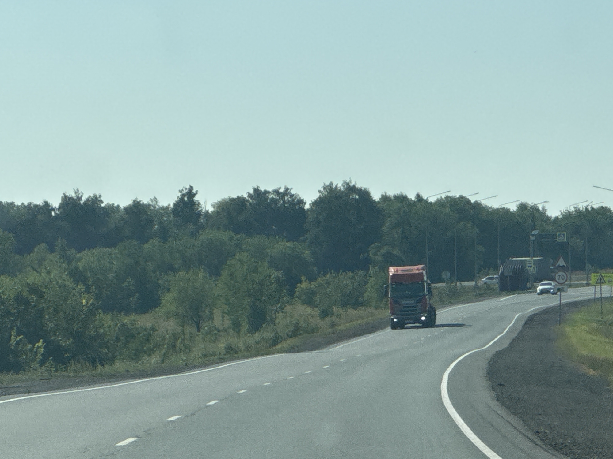 The image depicts a view from a vehicle traveling on a highway with dense green trees lining both sides of the road. The sky is a clear, pale blue. In the foreground, a grey asphalt road curves to the right, marked with white dashed lines. A red semi-truck is moving forward on the road in the mid-ground, and to its right, a white car is also traveling in the same direction. Further ahead, other vehicles are visible, including a dark-colored truck. Road signs are present, one indicating a speed limit of 70. The overall scene suggests a sunny day on a rural highway in Russia, likely near Perevolotsky, given the context.
