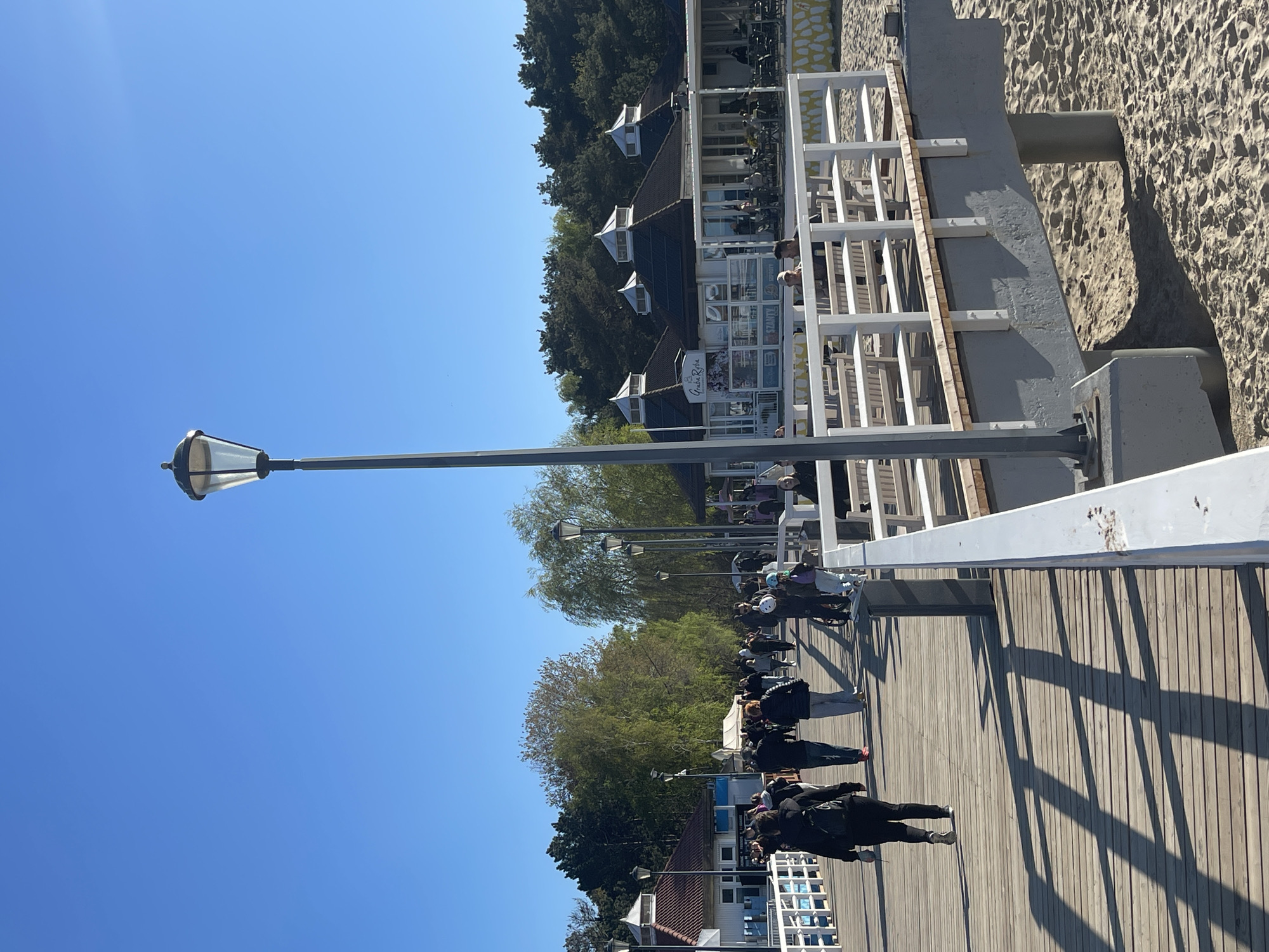 The image shows a wooden pier in Gdańsk, Poland, with a clear blue sky above. The pier is lined with streetlights and white railings, and several people are walking along it. On one side of the pier is a building with a restaurant called "Gruba Ryba" and the beach with sand. The pier is a popular tourist destination.