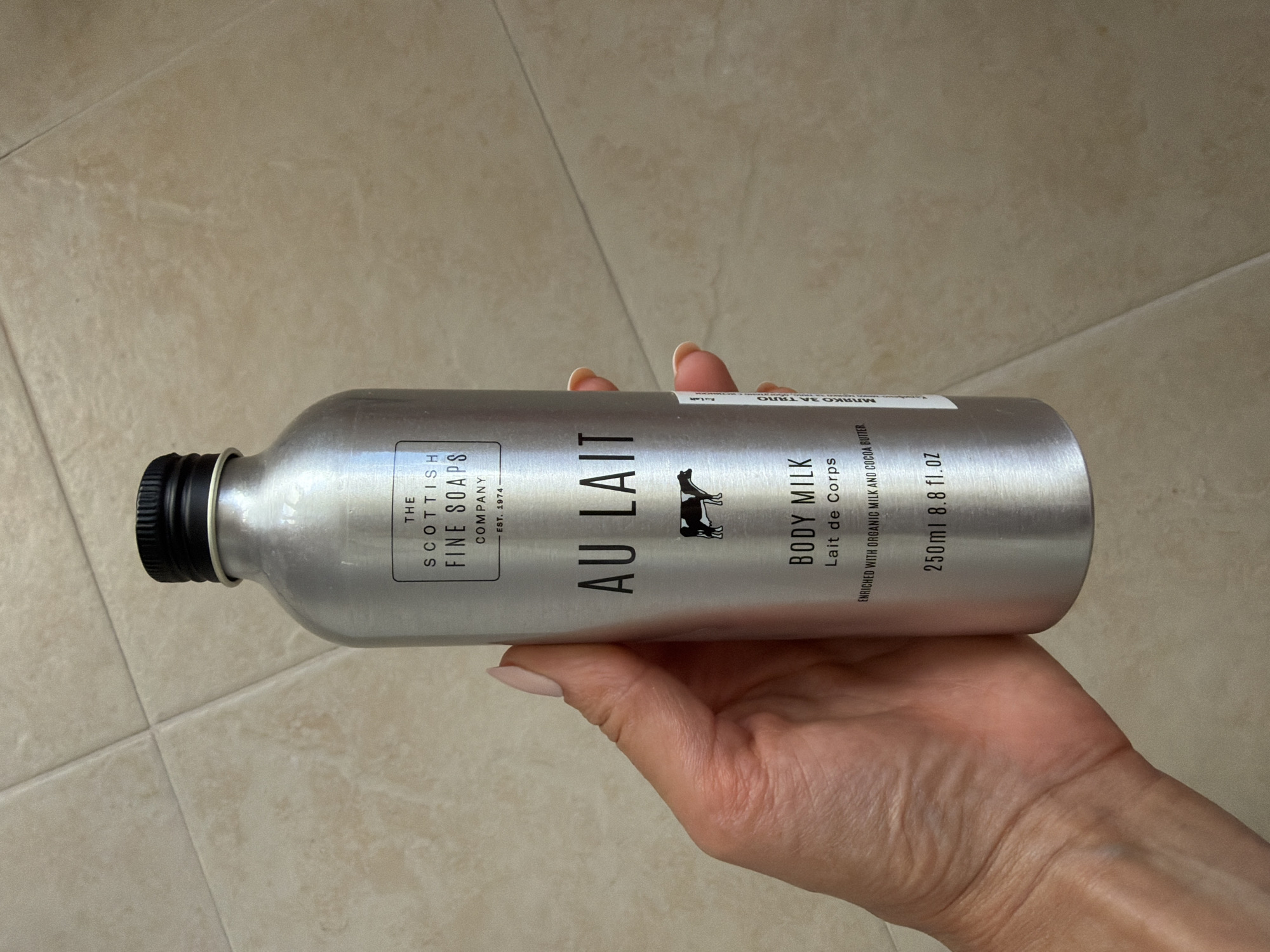 A hand with light skin and a light pink nail polish is holding a silver aluminum bottle of "AU LAIT BODY MILK" produced by "THE SCOTTISH FINE SOAPS COMPANY". The bottle has a black cap and features text and a small black and white illustration of a cow. A small white sticker with black text is attached to the right side of the bottle. The background consists of beige tiled flooring. The image was taken outdoors in Pazardzhik, Bulgaria.
