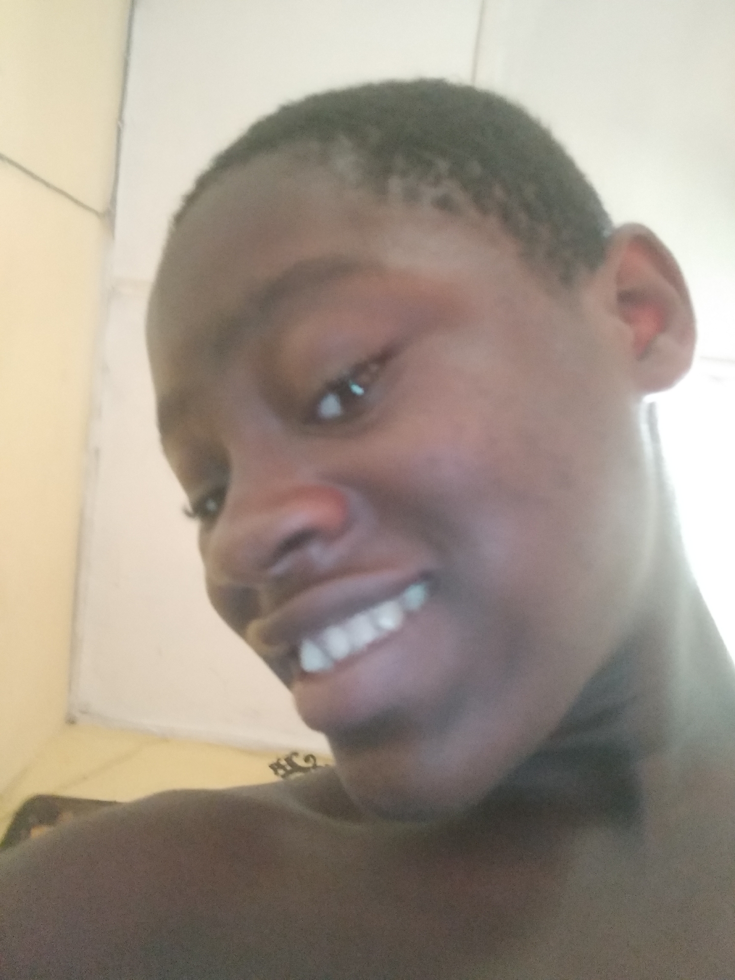 A close-up portrait of a young, dark-skinned person with short, dark hair, facing left, and smiling. The person's teeth are visible. The background consists of a partially visible off-white wall and a light-colored surface. The location is Shangisha, Nigeria.