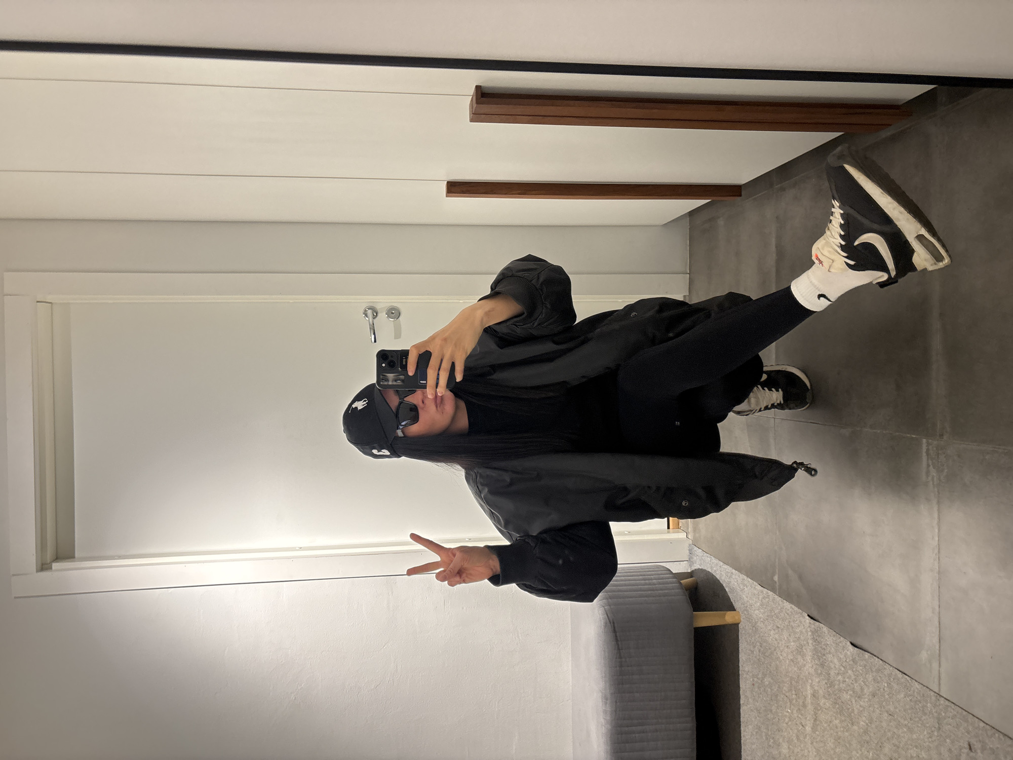 A person in dark clothing, including a jacket, leggings, and a cap with a logo, is taking a selfie in front of a mirror. They are indoors, possibly in a hallway or fitting room. The person is holding a cell phone in their right hand and making a peace sign with their left hand. They are wearing sunglasses. The background features a white door, gray flooring, and a gray, cushioned bench or seat. There are wood-colored elements on the wall behind the person as well. The location is unknown.