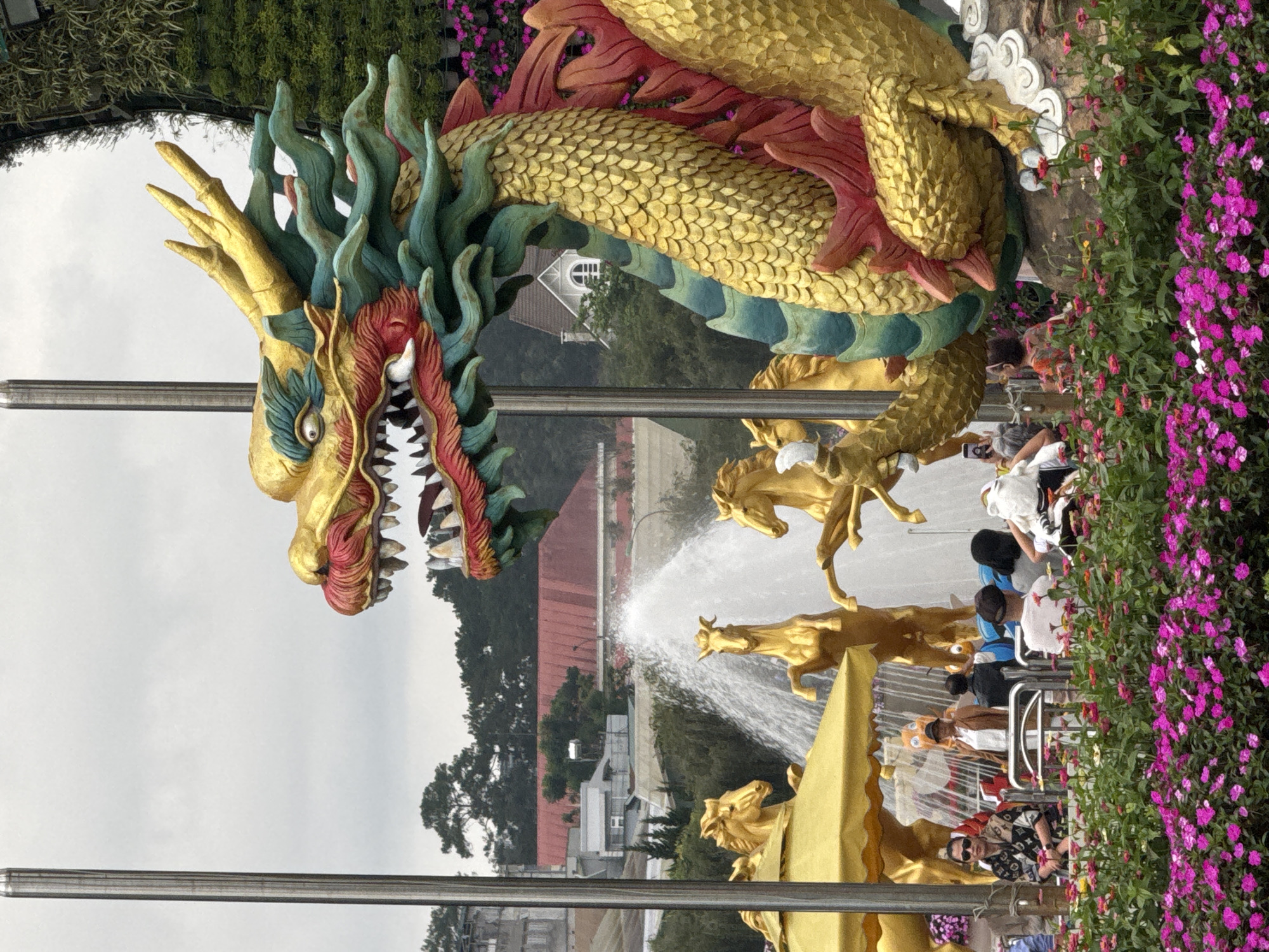 The image depicts a large, ornate golden dragon statue with turquoise and red accents, positioned outdoors. The dragon's head is in the foreground, its mouth open, revealing sharp teeth. Behind the dragon, there is a fountain with several golden horse statues. In the mid-ground, a crowd of people are visible, some seated and others standing, with a woman in a brown jacket and hat standing prominently. A bright yellow umbrella is situated near the fountain. The background shows buildings and trees under a cloudy sky. The foreground is filled with vibrant pink flowers. The scene is located in Phường Xuân Hương, Đà Lạt, Vietnam.