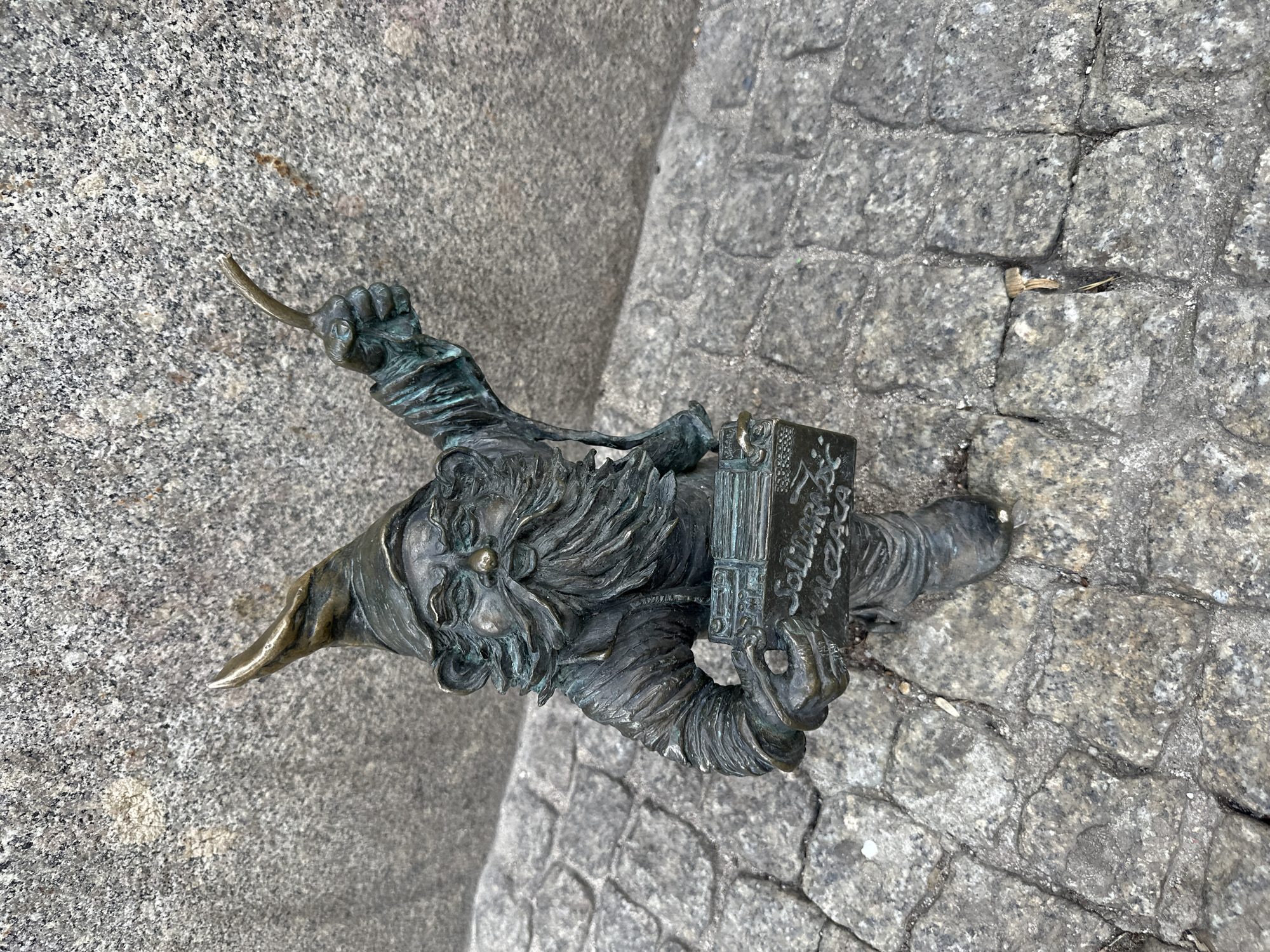 A bronze statue of a gnome stands on a cobblestone street. The gnome wears a pointed hat and has a long beard. He holds a book in his left hand and a pickaxe in his right. The book has inscriptions in Polish that read "Solidarnosc". The statue is in Wrocław, Poland, where these statues are common and a symbol of the city.
