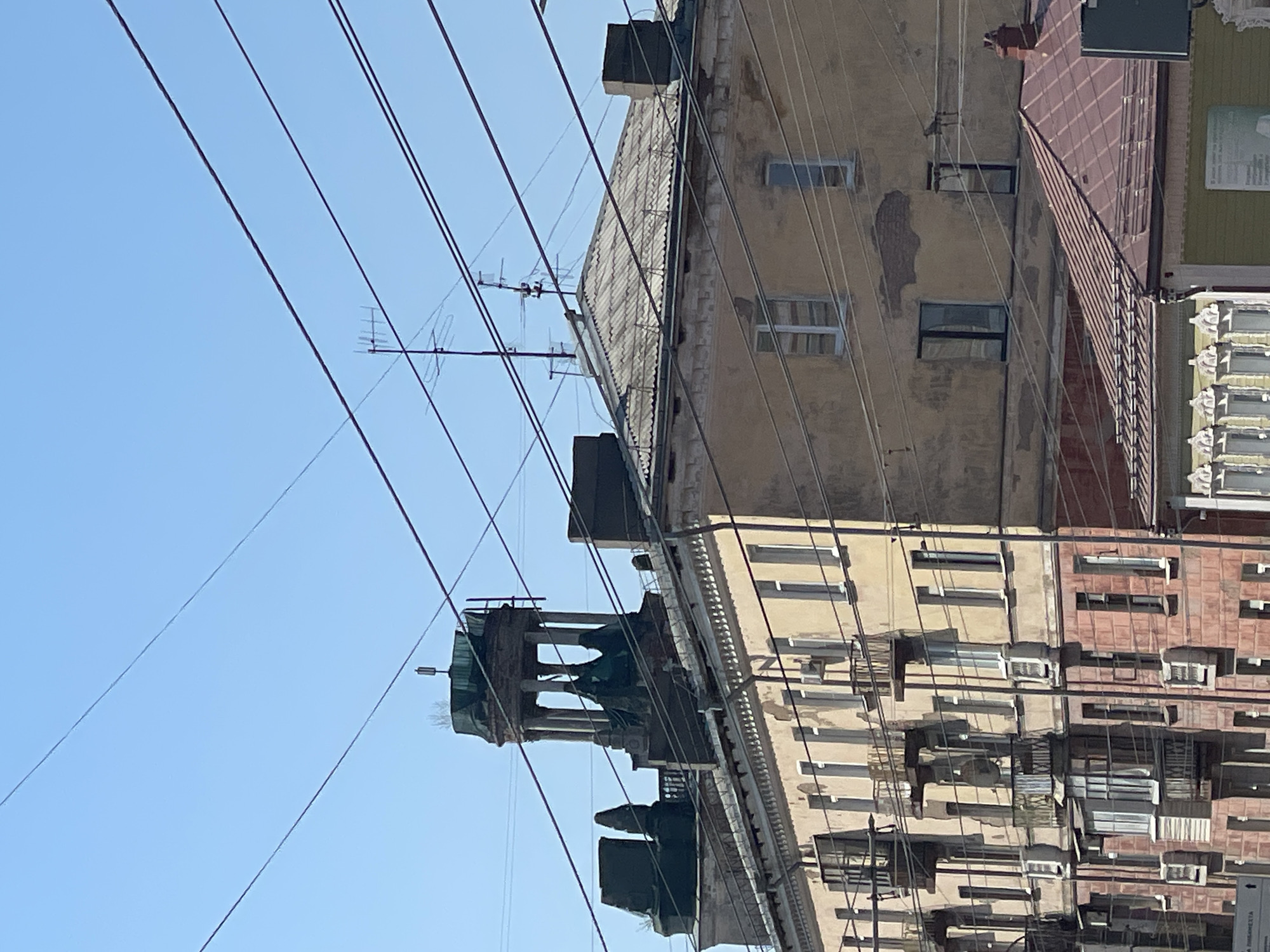 The image shows a building facade in Izhevsk, Russia, beneath a clear blue sky crisscrossed by numerous power lines. The building consists of multiple sections with varying architectural styles and degrees of disrepair. The section closest to the viewer has a brick facade with rows of windows, some with air conditioning units. A taller, yellowish section displays peeling paint and visible water damage. Above this is a roof with an antenna. A distinctive architectural feature, possibly a tower or cupola with open arches, is partially visible against the blue sky.