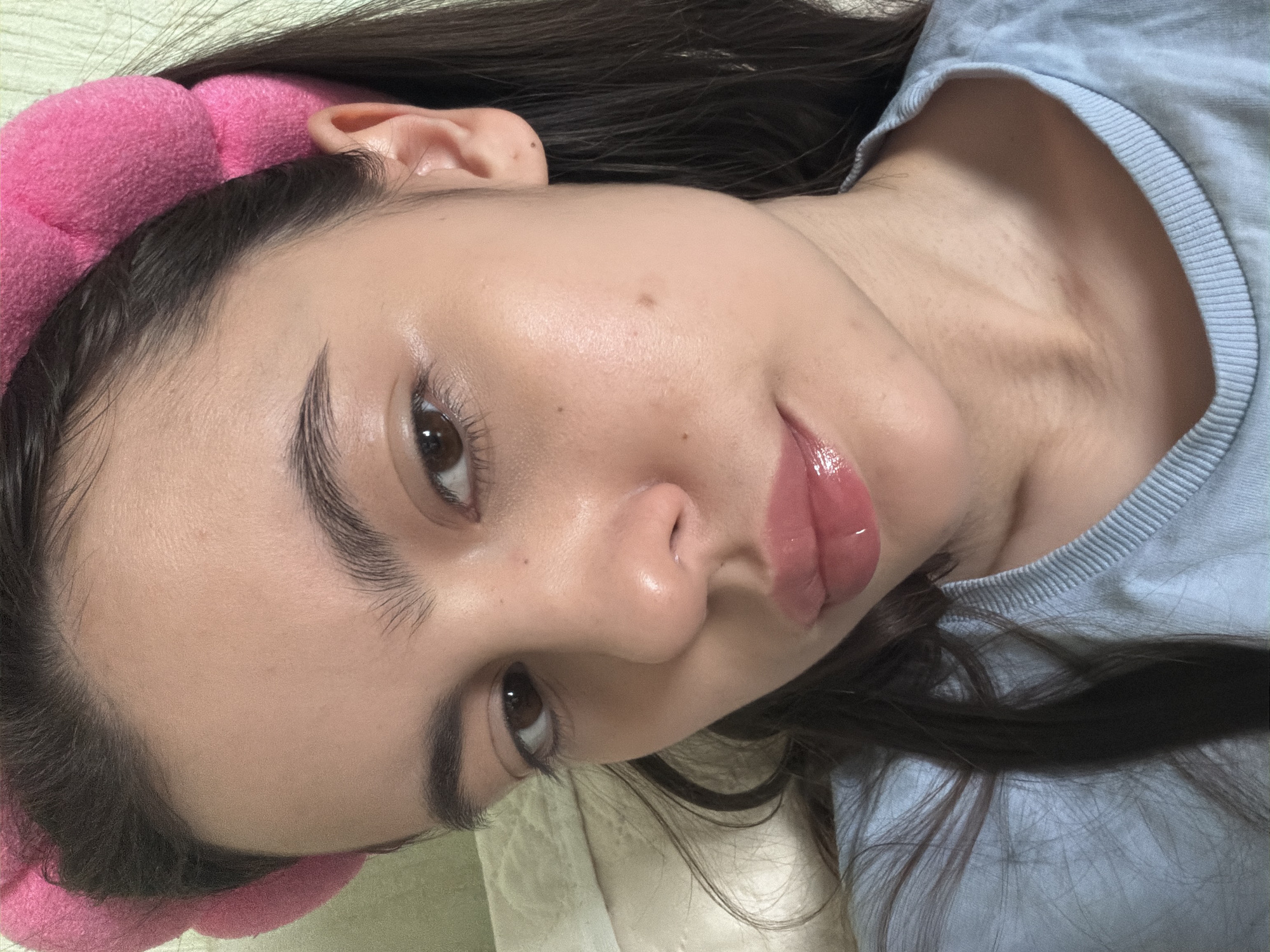 A close-up, slightly angled shot focuses on a young woman's face. She is wearing a pink, padded headband. Her dark hair is visible around the headband. She has brown eyes and fair skin with a glossy lip. She is wearing a light blue shirt. The background appears to be a white, textured surface, possibly a bedsheet. There's no information available in the image to identify the location as Chaikovsky, Russia.