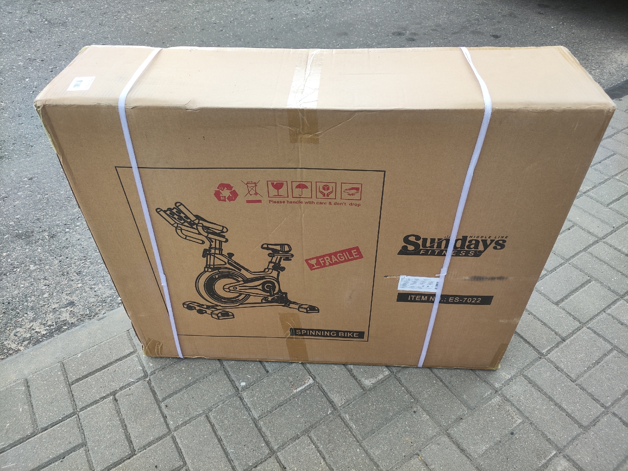 A large cardboard box containing a "SPINNING BIKE" from "Sundays FITNESS" is resting on a paved walkway. The box is secured with white plastic strapping. Various handling symbols, including "Please handle with care & don't drop" and a "FRAGILE" sticker, are printed on the box. The box has some wear and tear, with tape visible on the seams. The background appears to be an outdoor setting with asphalt and brick paving. The image is taken from a slightly elevated angle, looking down at the box. The item number "ES-7022" is clearly visible. There are no people visible in the image. The lighting suggests it might be daytime.