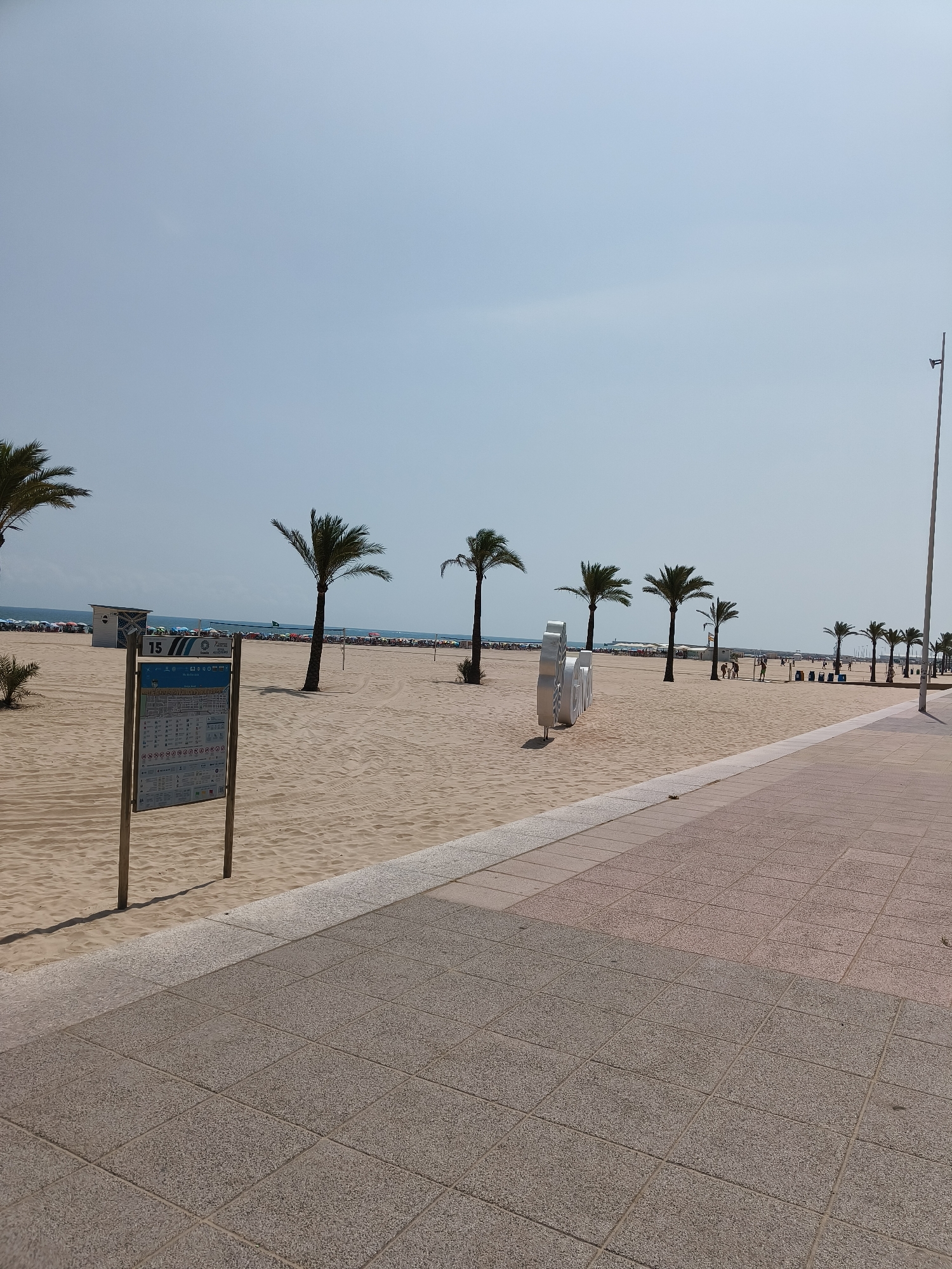 The image shows a wide, sandy beach in Gandia, Spain, on a clear day. A paved promenade runs along the right side of the scene, transitioning from a lighter pinkish-tan to a darker grey. Several palm trees are scattered across the beach, with a line of them visible along the water's edge in the background.
In the foreground left, a large informational signpost with a map and various icons is planted in the sand. To the left of the signpost, a small, white beach hut with a dark roof is visible. Further back on the beach, numerous beach umbrellas and people are scattered, indicating a populated area.
In the mid-ground, a large, white, sculpted object with curved lines, possibly a decorative installation, stands on the sand. People are walking and congregating in this area. In the far background, the calm blue sea meets the horizon under a pale blue sky. A tall, thin lamppost is visible on the far right side of the image, extending towards the sky. The sand is marked with tire tracks and footprints, suggesting recent activity.
